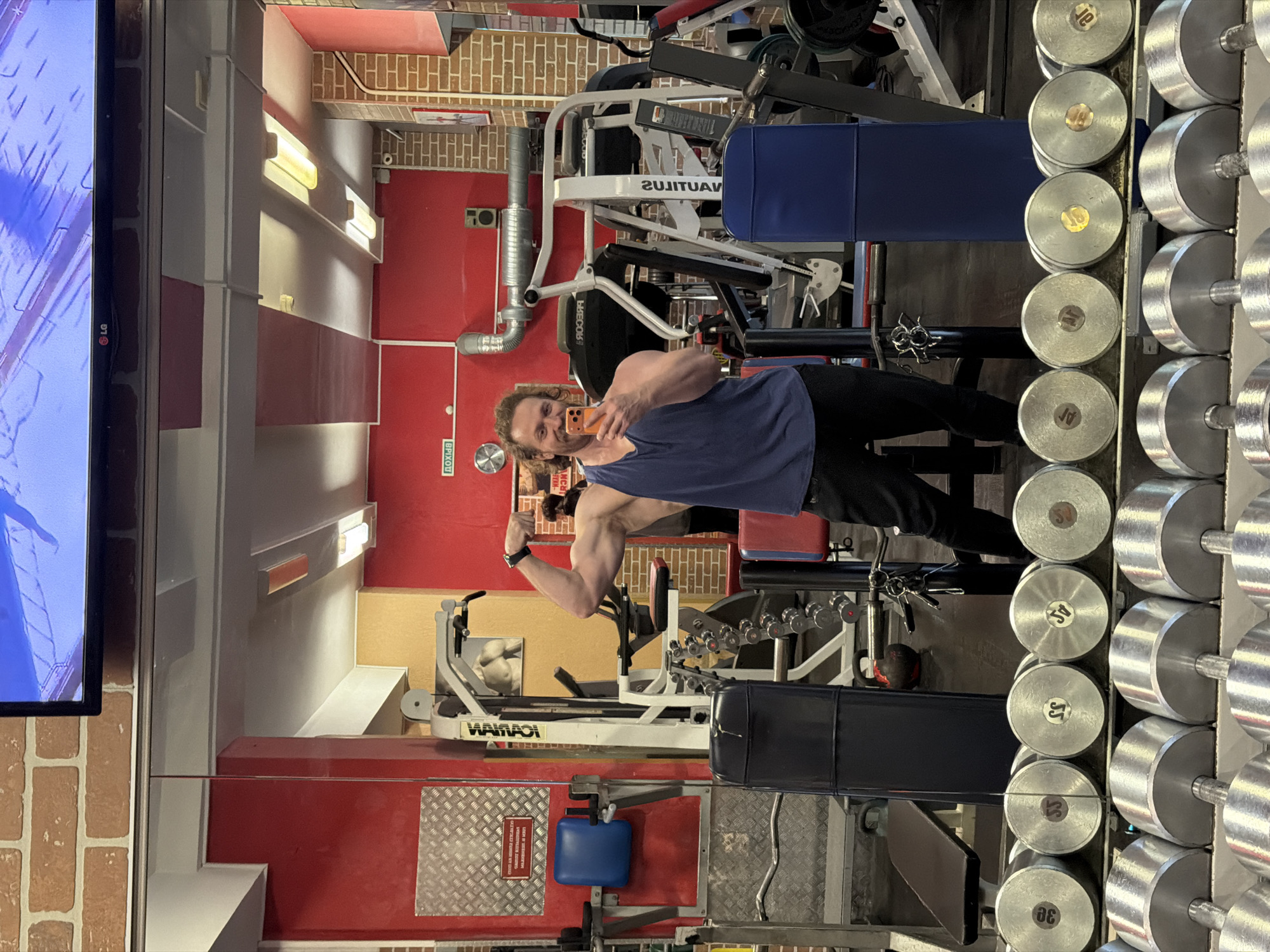 A man with long brown hair in a ponytail stands in a gym, holding a phone in his right hand to take a selfie, flexing his left bicep. He wears a blue sleeveless shirt and black pants. To his right, there is a rack of dumbbells. Behind him, various weightlifting machines are visible. The gym walls are a mix of red paint and brick. The city of Saint Petersburg, Russia, is not discernible from the image.