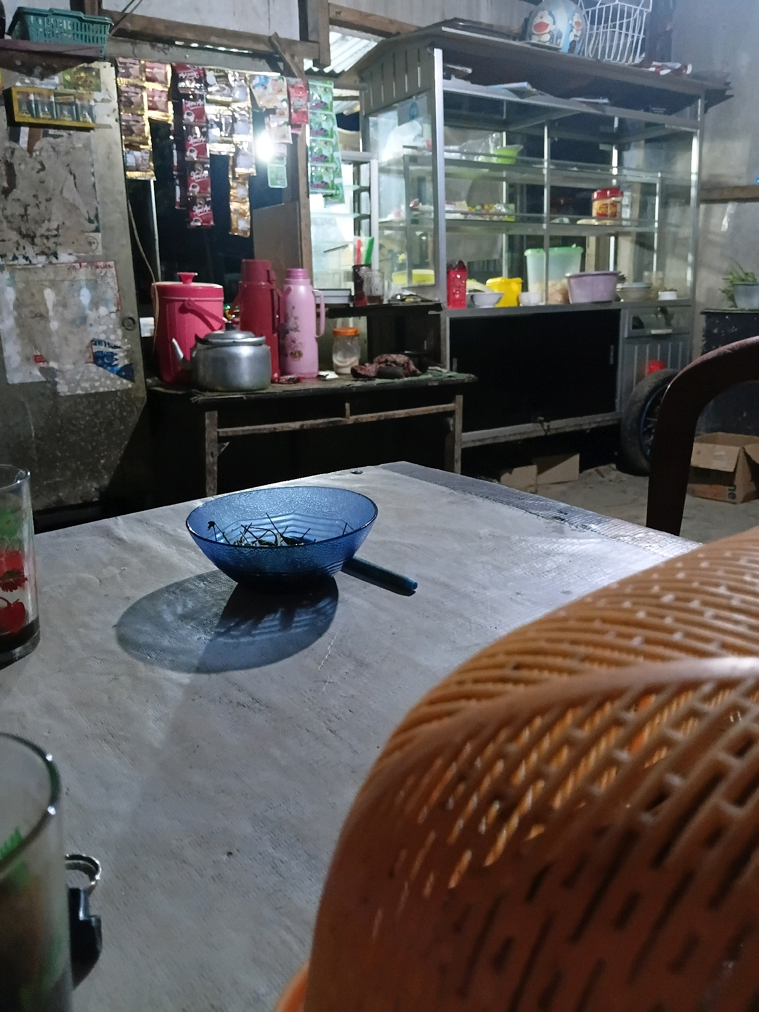 The image captures a dimly lit outdoor food stall or small eatery at night. The foreground features a weathered wooden table with a blue glass bowl containing some green stems. To the left, a glass with a fruity pattern holds a dark beverage. In the midground, a metal shelf unit displays various items, including two pink thermoses, a silver teapot, and packets of instant noodles or seasonings hanging on the wall. A glass of water is also visible. The background shows more shelves filled with containers and what appear to be food products, suggesting a retail or preparation area. The overall setting is cluttered and rustic, characteristic of a local, informal establishment. The presence of the wooden table, thermoses, and display shelves strongly implies a food and beverage service, likely in a residential or semi-commercial area. While the location is specified as Bantarbarang, Indonesia, there are no explicit visual cues within the image itself to confirm this specific geographic detail.