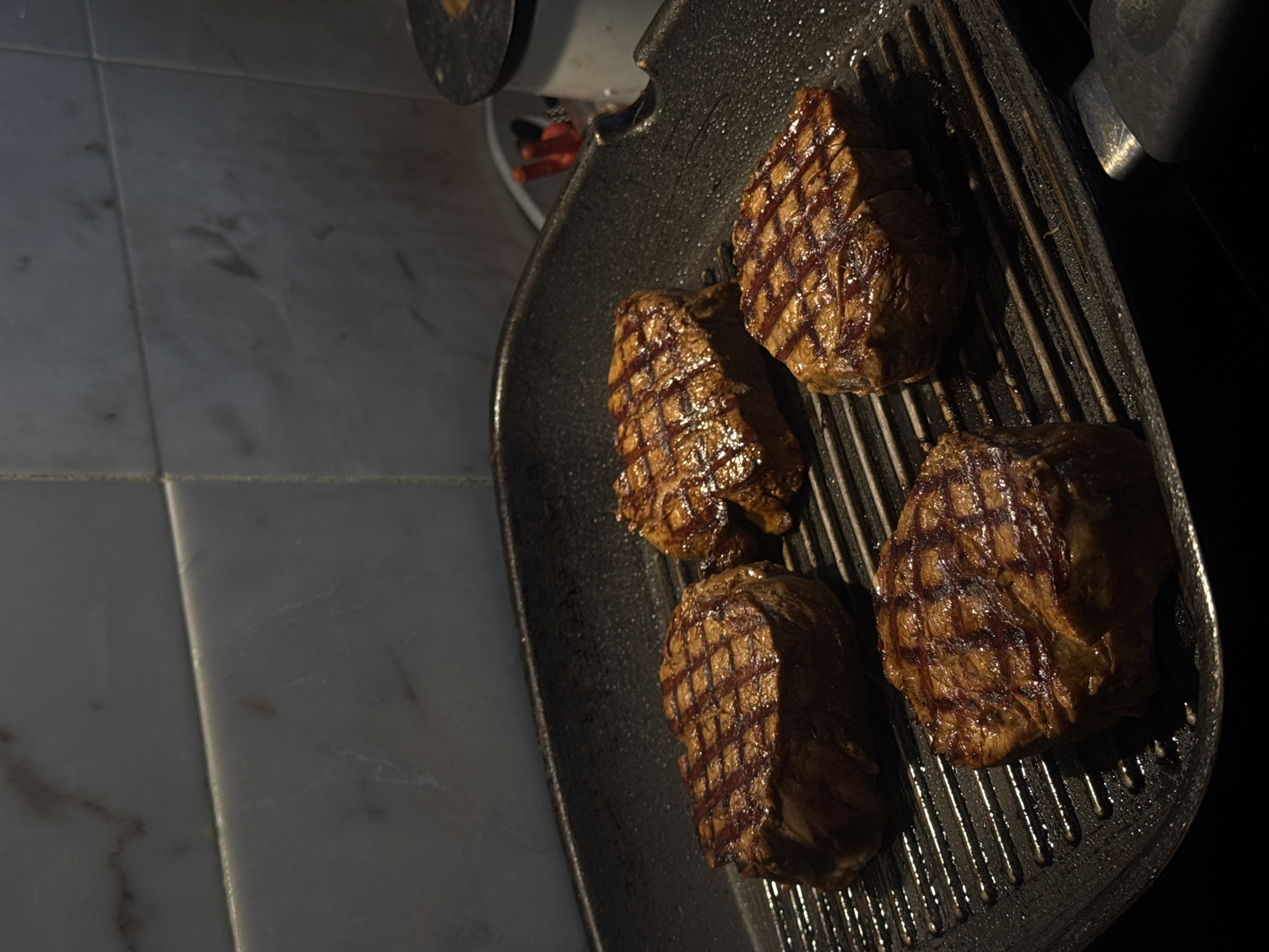The image shows four pieces of cooked meat, likely steaks, in a dark gray grill pan. The meat has distinctive grill marks. The grill pan appears to be on a stovetop. Part of the stove and a tiled surface are visible. The tiling is light colored. It is unknown if the location is Ptichnoye, Russia. No people, specific subjects, interactions, or environment details are observable beyond what is described.