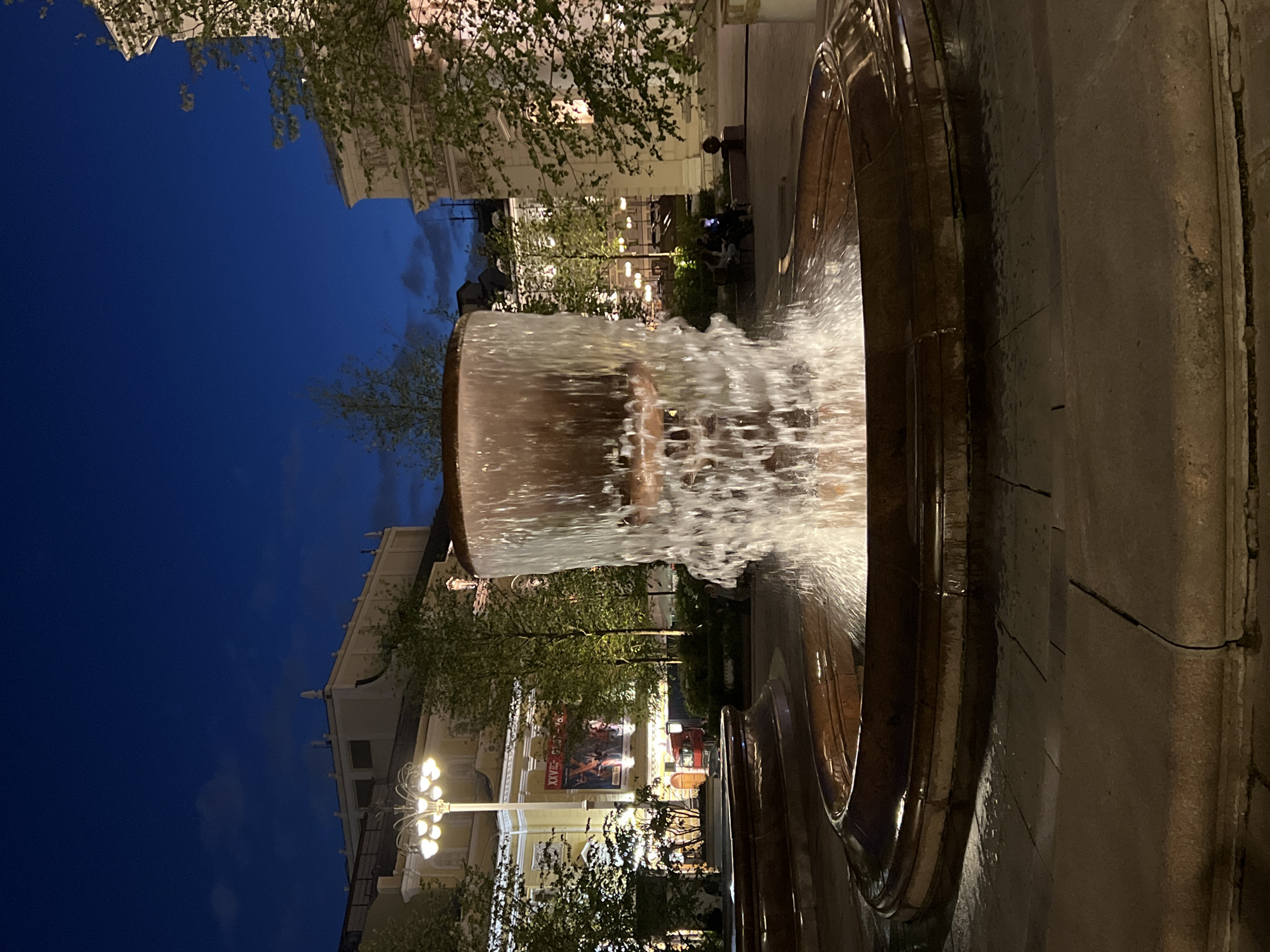 A multi-tiered fountain with water cascading down its sides is the central focus of the image, set against a twilight sky of deep blue with hints of clouds. The fountain basin is constructed from reddish-brown stone with a polished rim, and the surrounding ground is paved with stone slabs.
Behind the fountain, trees with sparse green foliage are visible, illuminated by streetlights. In the background, buildings with light-colored facades and architectural details stand under the night sky. One building features a large poster with text in a foreign language and images of people, suggesting it might be a theater or cultural venue. A streetlamp with multiple glowing bulbs is positioned to the left of the fountain.
Several people are casually seated on benches in the mid-ground, some looking at their phones. The overall scene suggests an outdoor urban public space, likely a square or plaza, at dusk or early evening. The lighting creates a moody and serene atmosphere.