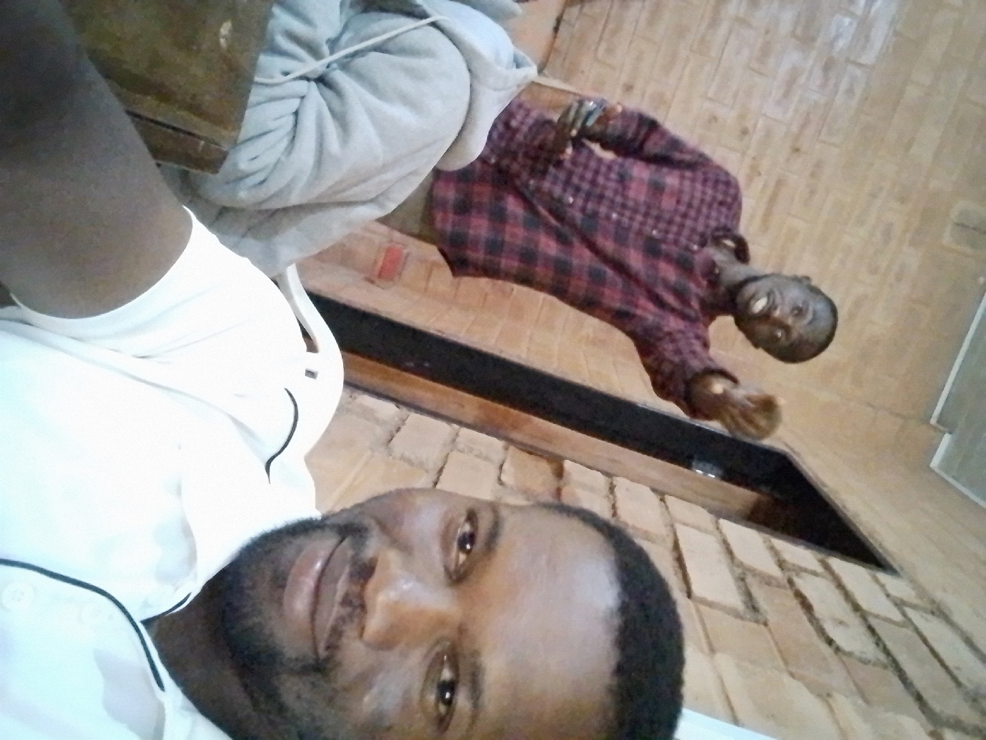 Two men are in an indoor setting with a brick wall. The man in the foreground is facing the camera and smiling. He has dark skin and a beard, and is wearing a white collared shirt. The man in the background is wearing a red and black plaid shirt and is pointing his finger towards the camera. He is looking to the side and appears to be speaking. There is a wooden table in front of the man in the foreground.