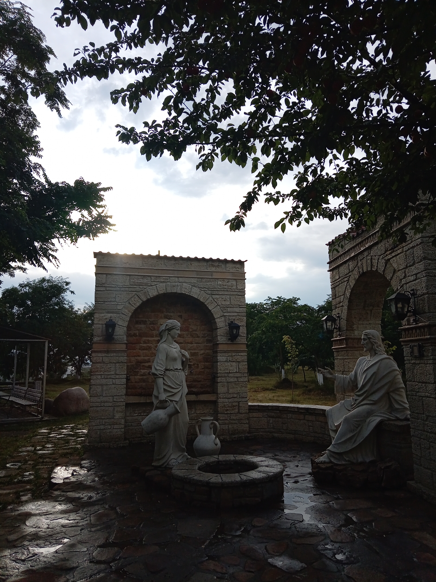 This image depicts a sculpture garden with two statues, one of a woman carrying a water jug and the other of a seated figure with outstretched hands, likely representing Jesus. The statues are situated in front of stone structures with arched openings. The ground is paved with irregular stones, and there are well-maintained plants and trees in the background. The sky is overcast with some hints of sunlight breaking through. This scene is located in Yessentuki, Russia, likely a tourist attraction or a place of worship, given the artistic and religious themes of the sculptures and the overall ambiance of the garden.