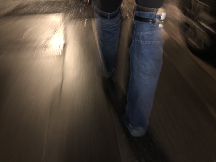 A blurry, low-angle shot captures two legs clad in blue denim pants or jeans, each secured with a black belt featuring a silver buckle. The lower portion of the frame shows a dark, reflective surface, possibly pavement, streaked with blurred light and shadows. The background is heavily blurred, creating a sense of motion. The overall lighting is dim, with the bright streak of light adding an element of mystery.