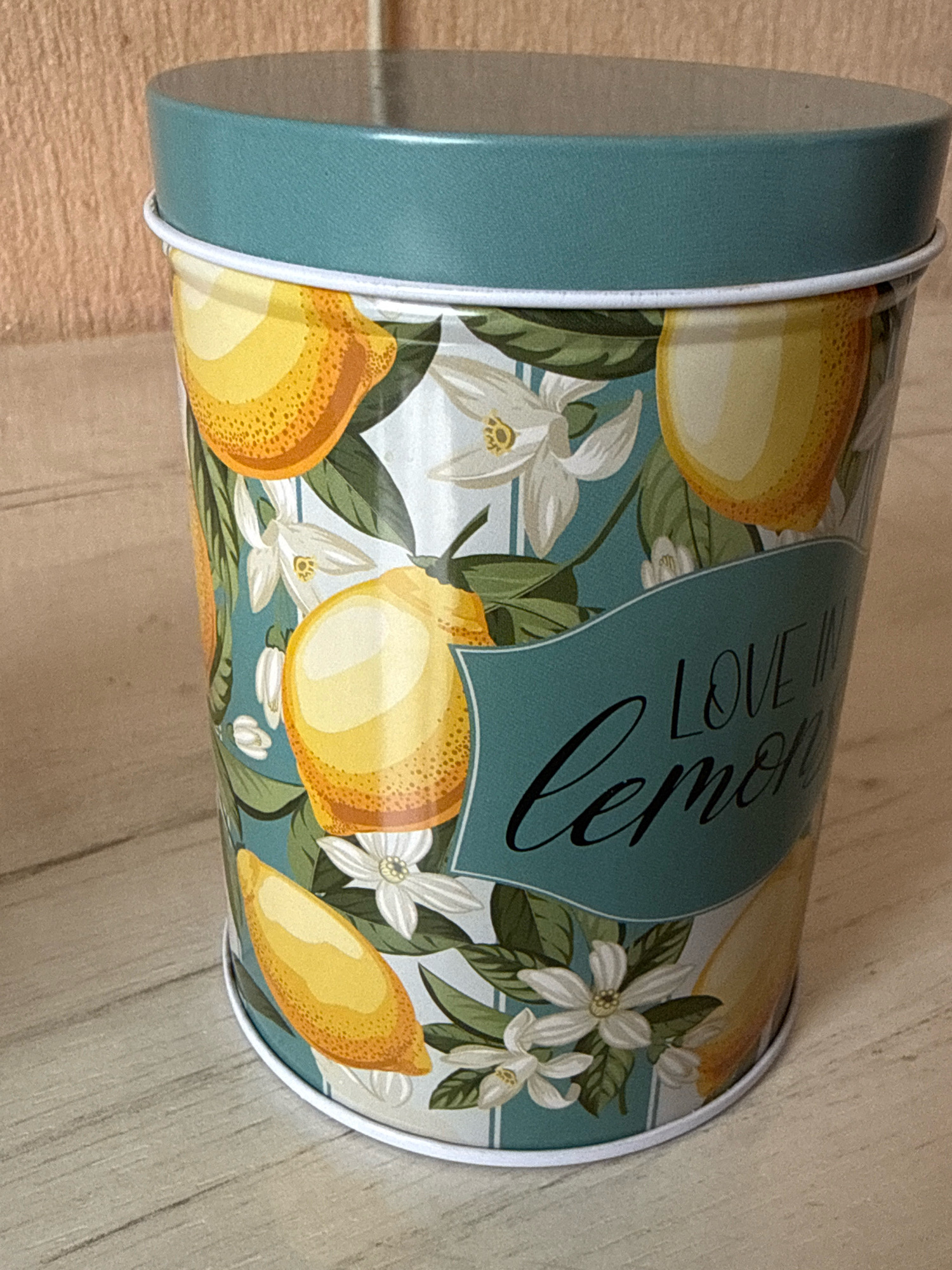 A tall, cylindrical tin can with a teal lid is shown against a light-colored wooden surface. The tin can is decorated with a pattern of lemons and white flowers on a light blue background. A dark teal banner across the can displays the words "LOVE IN Lemon" in a cursive font.