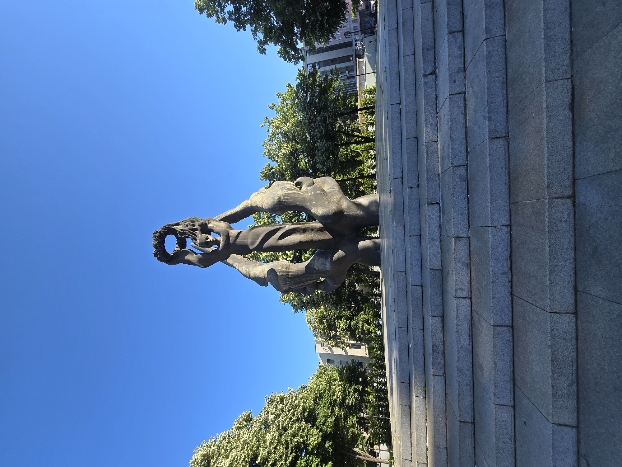 A bronze statue stands atop a tiered gray stone structure, set against a clear blue sky. The statue appears to depict figures intertwined, with one holding a wreath. Trees and buildings are visible in the background.