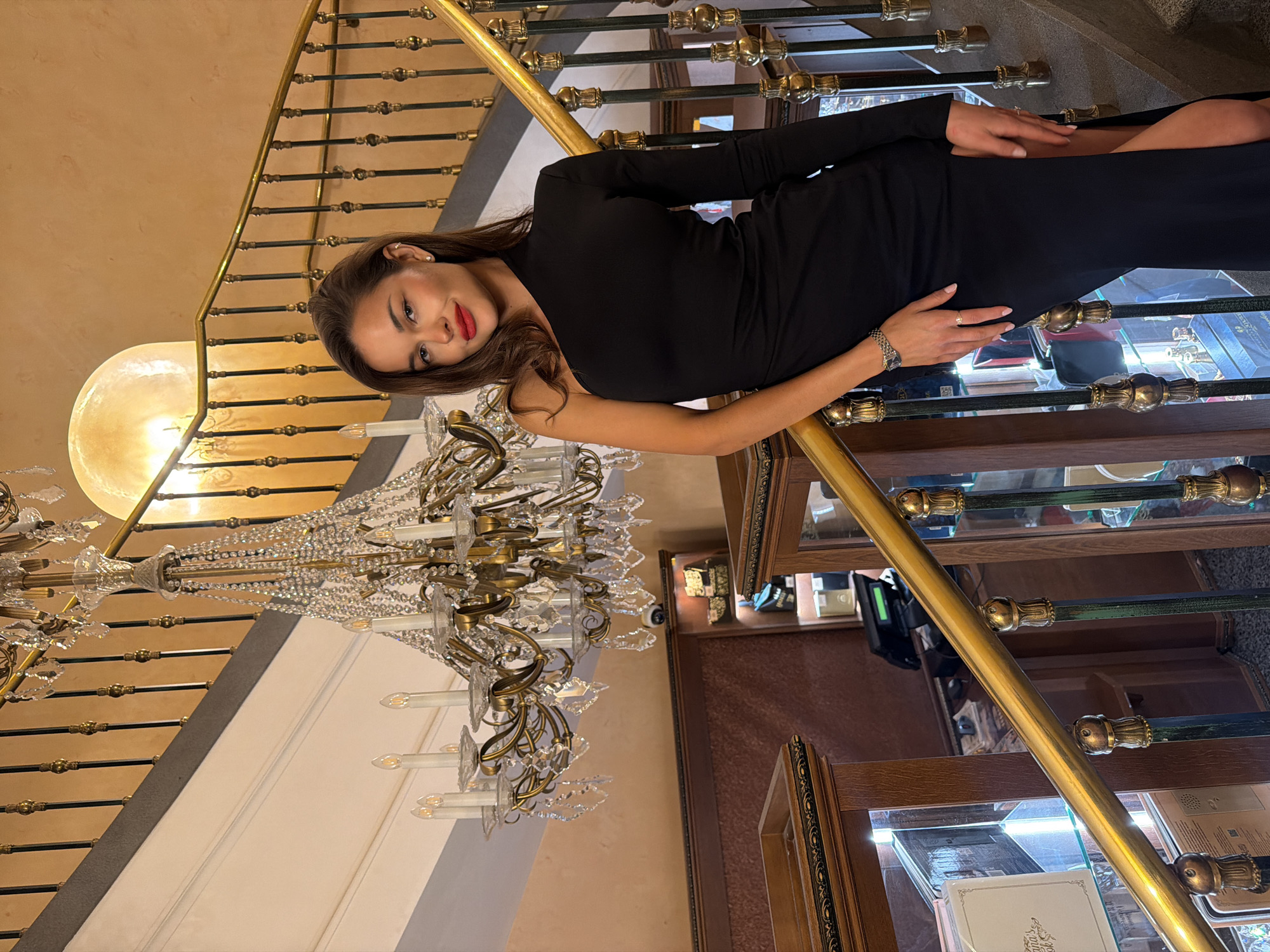 A young woman is captured posing on a grand, ornate staircase within an opulent indoor setting. She stands, leaning casually against a polished golden handrail, looking directly at the camera with a subtle smile. She has long, dark brown hair and is wearing a form-fitting black dress with long sleeves. Her facial features are highlighted by red lipstick and light makeup. A silver-toned watch is visible on her left wrist, and a small stud earring adorns her left ear.
The staircase features richly detailed golden balusters and a dark, possibly wooden or marble, surface. A large, elaborate crystal chandelier, adorned with numerous candle-style lights, hangs prominently from the ceiling above the stairs, illuminating the scene. Another large, circular light fixture casts a warm glow in the upper left background. To the right and below the staircase, several ornate wooden display cases with glass fronts are visible, some internally lit, suggesting they hold merchandise. The walls are a warm, light color, complementing the luxurious fixtures. The overall ambiance is one of sophistication and elegance, indicative of a high-end establishment in Moscow, Russia. There is no visible text in the image.
