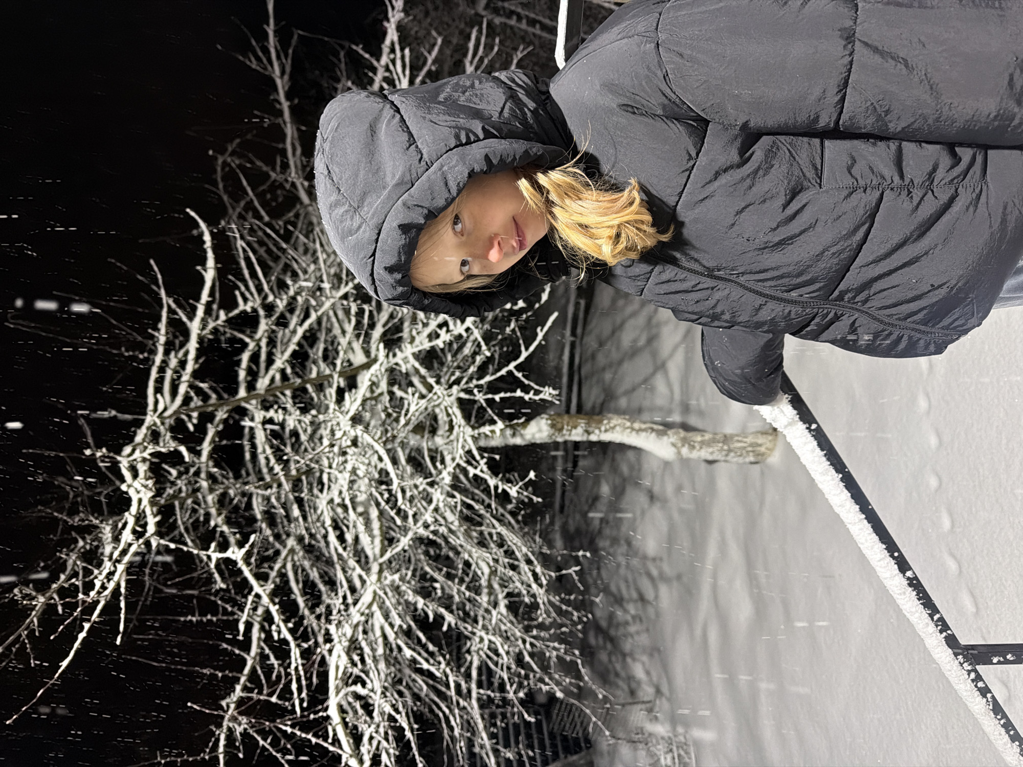 A nighttime photograph, rotated 90 degrees counter-clockwise, depicts a person in a black hooded jacket against a snowy backdrop. The person's face is visible, with blonde hair partially visible under the hood. To the left of the person, a snow-covered tree with many branches stands out against the dark sky. Snowfall appears throughout the scene. In the lower right corner, a snow-covered railing is visible. The location is purportedly Markovo, Russia, based on additional information, though this cannot be confirmed from the image alone.