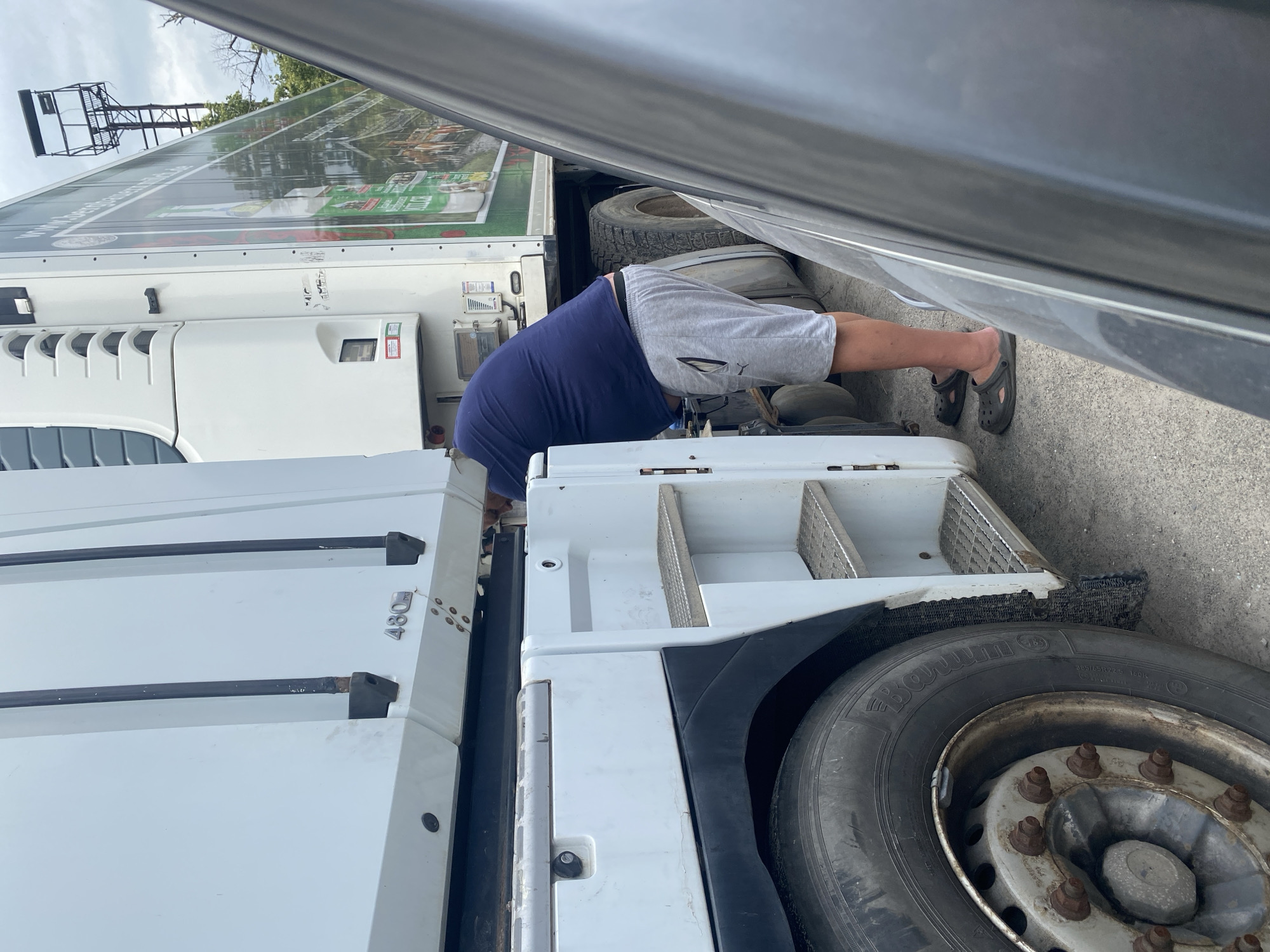 A man wearing a blue t-shirt and grey shorts is bent over, appearing to be working on the side of a white semi-truck in Dnipro, Ukraine. The truck has a refrigerated trailer with advertising on its side. The man is wearing dark clogs. The truck is parked on an asphalt surface, and another dark-colored vehicle is partially visible in the foreground, obscuring part of the scene. The sky is overcast.