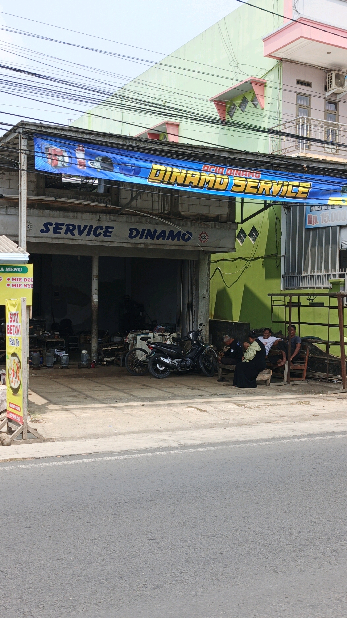 The image captures a street-level view of a commercial establishment in Desa Wonoharjo, Indonesia, likely a service shop. A prominent blue banner stretches across the top of the building, featuring text that reads "ACID DINAMO SERVICE" in large, bold lettering with a yellow outline. Beneath this banner, the building's facade displays the words "SERVICE DINAMO" in blue paint. The shop entrance is open, revealing a dimly lit interior with various equipment and machinery, including what appear to be motor parts and a bicycle.
In front of the shop, three men are seated on wooden benches, engaged in conversation. One man, wearing a black outfit, is partially obscured. The other two are casually dressed. A black motorcycle is parked in front of the entrance, leaning on its side stand. To the left of the entrance, a vertical yellow sign with red and white text, possibly a menu or advertisement, is propped up by wooden supports. The surrounding environment is a paved street with a concrete sidewalk in front of the building. Power lines crisscross the sky above. The building itself has a light green painted exterior on the right side, with architectural details including windows and a balcony. The overall impression is that of a bustling commercial area, with daily life unfolding outside the service shop.