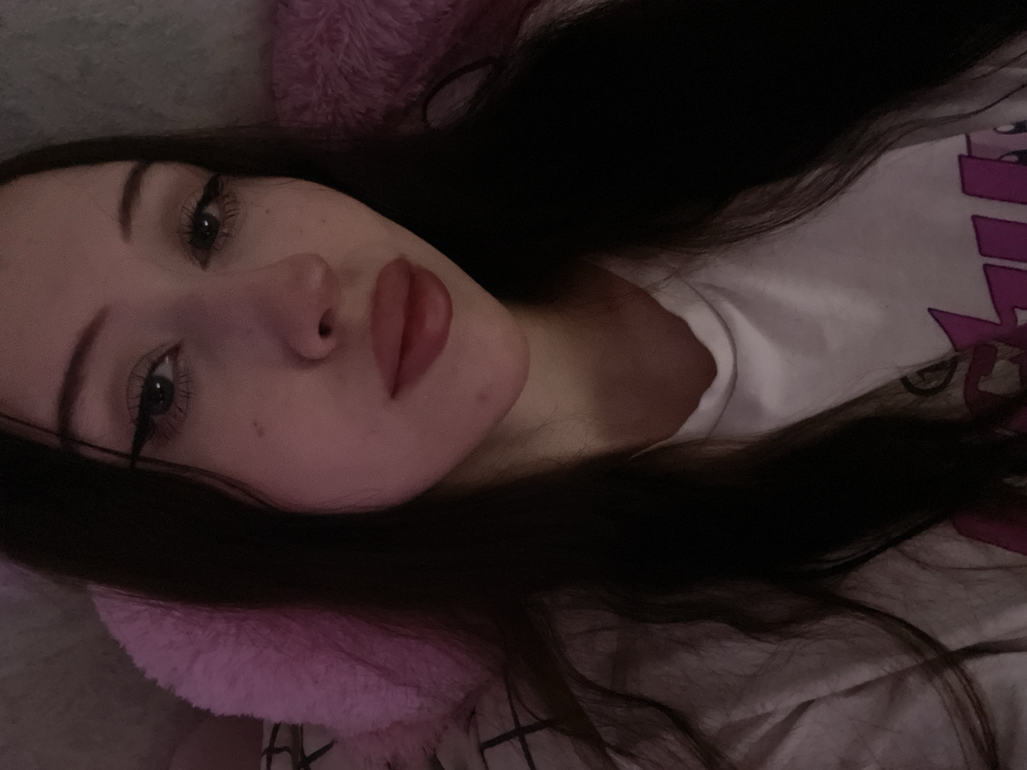 The media file is a close-up, low-angle, selfie-style photograph of a young person with long, dark hair lying down. The subject is wearing a white t-shirt with a visible graphic print on the right side, featuring purple lettering or shapes. The person has their eyes open, with notable eye makeup including winged eyeliner and mascara. Their lips are slightly parted and appear to be wearing lipstick. The lighting is dim, suggesting it might be nighttime or an indoor setting with low light. The background includes soft, fluffy pink material, possibly a pillow or blanket, and a darker surface that could be bedding or furniture. The overall mood of the image is intimate and relaxed. There are no clear indicators of the time of day or weather, nor specific location cues beyond the provided context of Votkinsk, Russia. The subject's expression is neutral, with a slight gaze towards the camera. No other individuals or distinct objects are prominently featured.