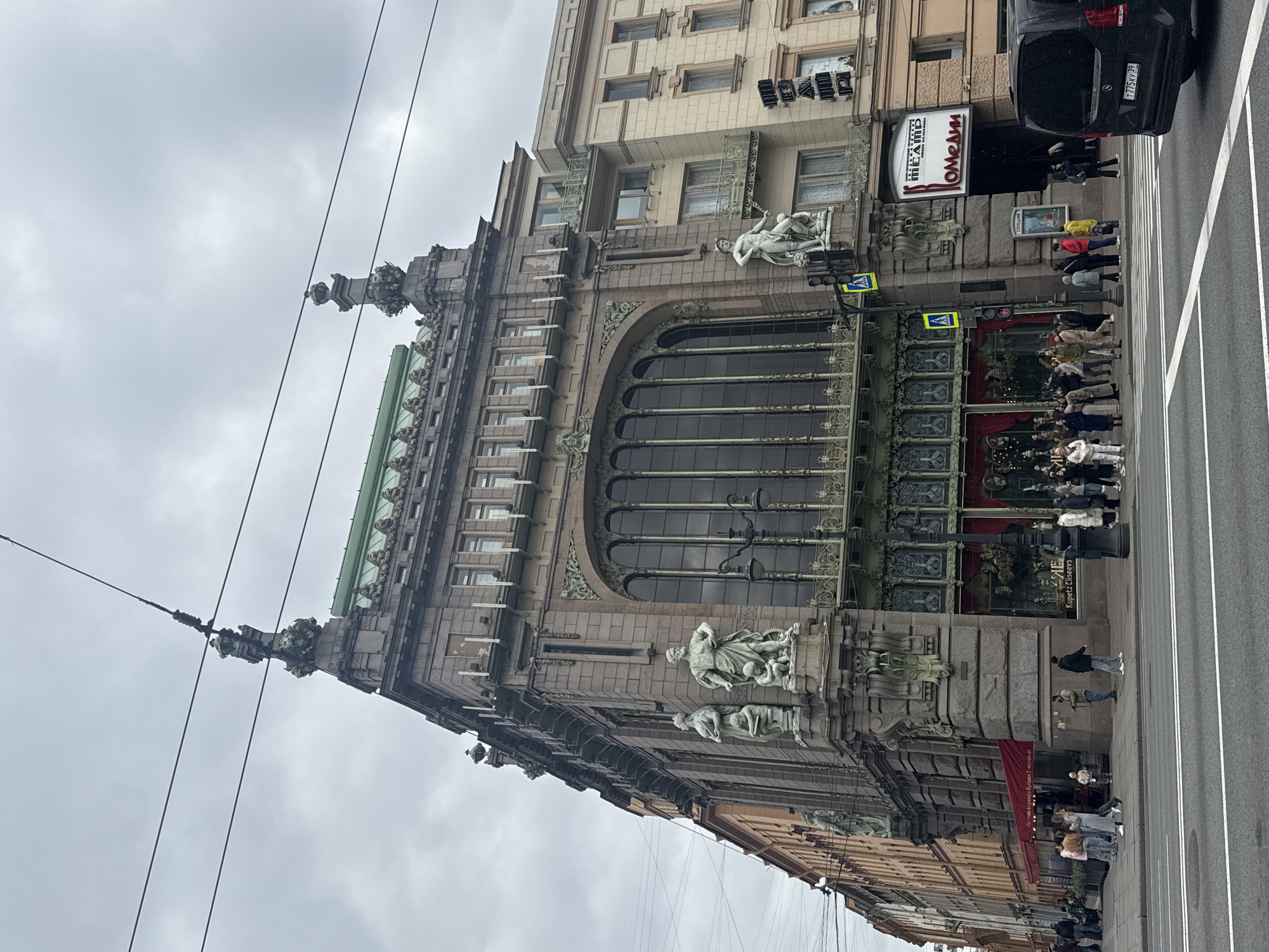 This image depicts the Eliseev Emporium, an Art Nouveau building in Saint Petersburg, Russia, on a cloudy day. A large arched window dominates the façade, adorned with sculptures and intricate metalwork. Several people stand on the sidewalk near the entrance. A black van is visible on the road next to the building. Traffic lights and overhead power lines are also present.