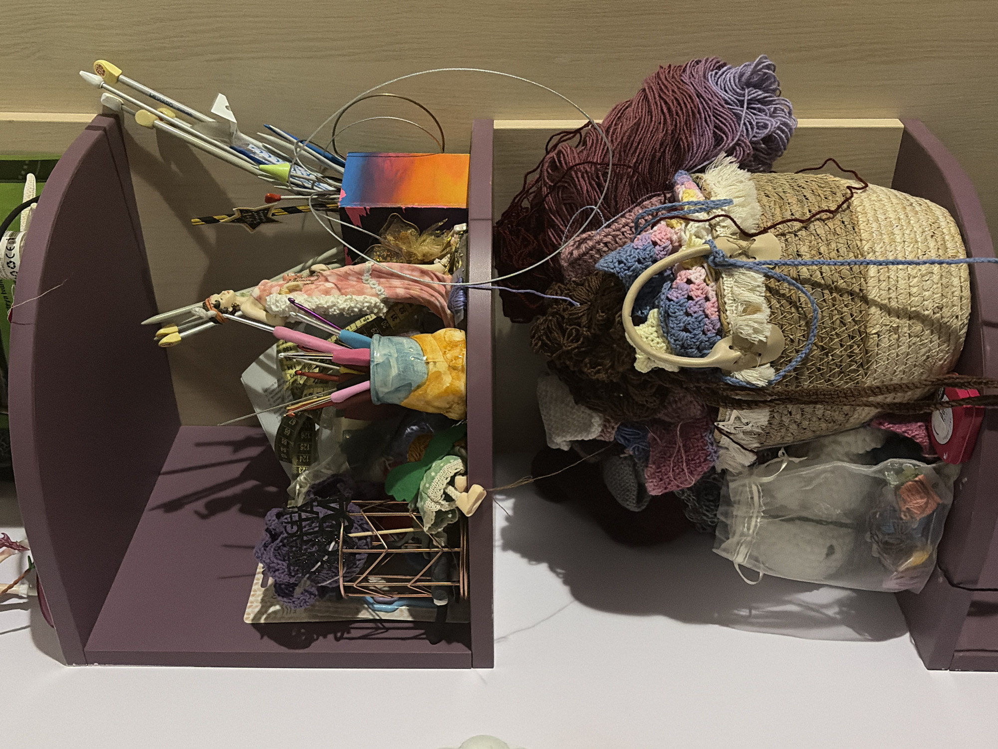 The image depicts a craft shelf filled with yarn, knitting needles, crochet hooks, and various other crafting supplies. The shelf is a deep purple color and is mounted on a white wall. On the top shelf, there are several knitting needles, a figurine of a woman in a pink dress, and a decorative container holding crochet hooks. There are also various balls and skeins of yarn in different colors, including purple, brown, and pink. On the bottom shelf, there is a large basket filled with yarn and some crochet projects. There is also a sheer drawstring bag containing smaller balls of yarn and other crafting items. A small red tin is also visible on the bottom shelf. The lighting suggests an indoor setting, possibly a home or a craft studio. There is no indication of the city of Vratsa, Bulgaria, in the image.