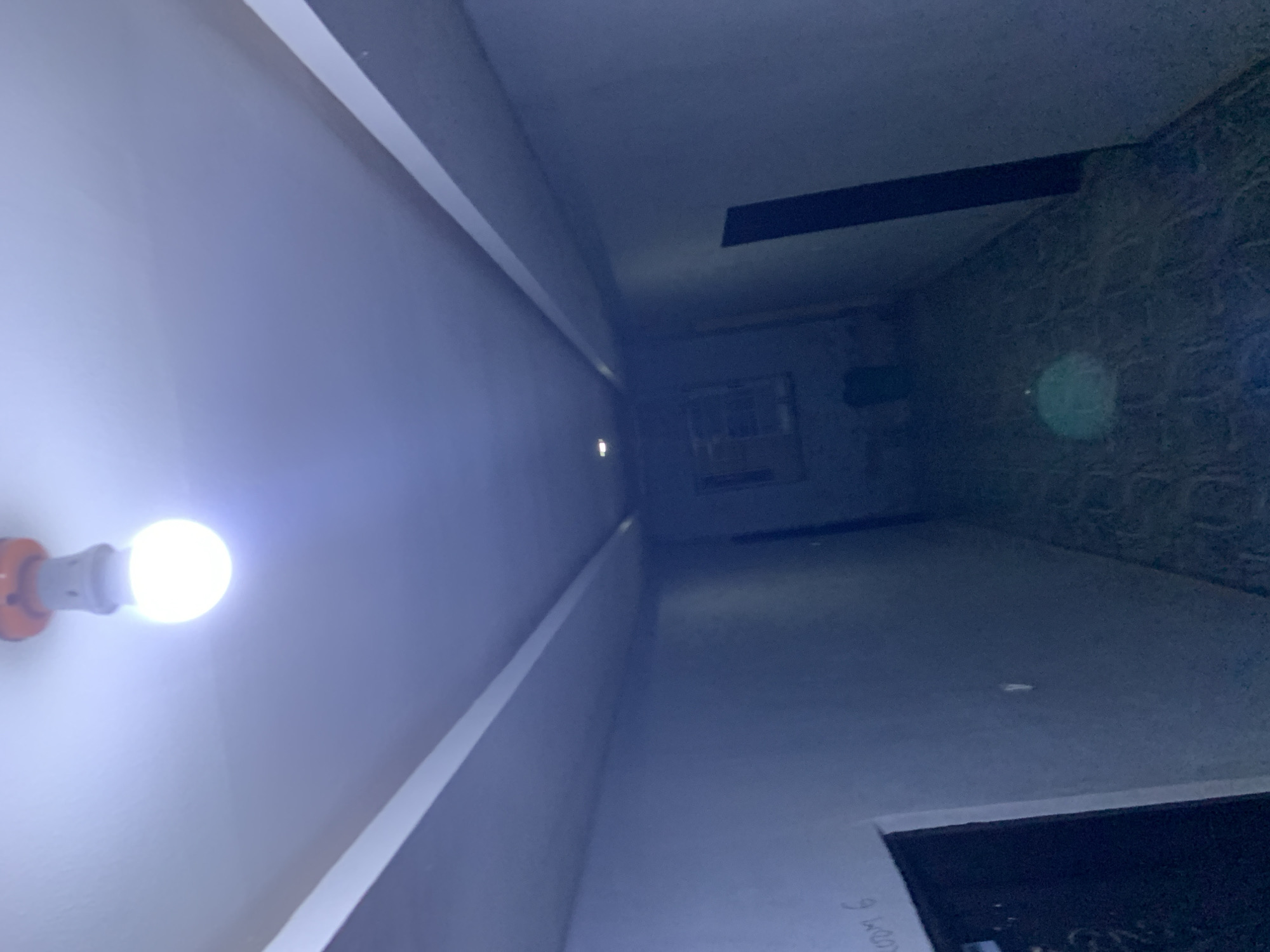 This image captures a view looking down a narrow, interior hallway or corridor, presumably located in Kabong, Nigeria. A single, bright, unshaded white spherical LED light bulb hangs from the ceiling in the foreground, casting strong illumination downwards and forward, which lends a prominent blueish tint to the entire scene.
The ceiling is a light, possibly off-white, surface featuring a symmetrical architectural element: two parallel, raised or recessed rectangular panels extend longitudinally along its length, framing a central flat section. The walls on both sides of the hallway are painted a dark, muted gray or blue-gray. On the left wall, in the foreground, the handwritten text "ROOM 6" is visible. To the left of this text, a dark, rectangular doorway is partially visible, indicating an entrance. Further down the corridor, another dark, vertical rectangular shape, likely a doorway, is visible on the right wall. At the far end of the hallway, a window with a visible grid or pane structure is present, but details beyond its outline are indistinct due to distance and dimness.
The floor is covered with a dark, textured material, possibly tiles, which shows faint patterns and reflections. A circular, light green lens flare is distinctly visible on the floor in the lower central portion of the image. The hallway appears unoccupied, with no people, discernible objects (other than architectural features), or actions taking place. The scene conveys a functional, artificially lit interior space.