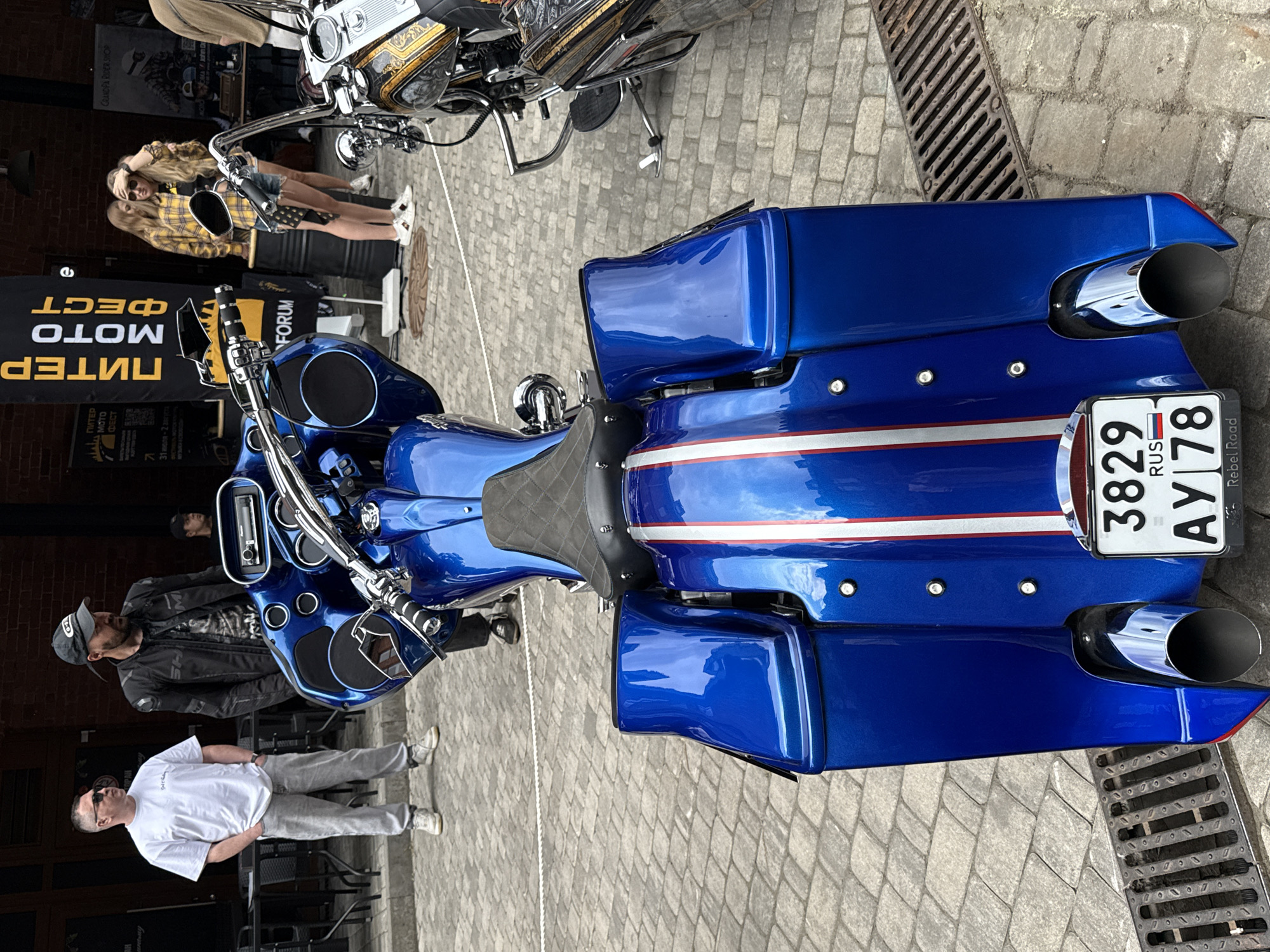 The image shows a blue motorcycle parked on a cobblestone street in Saint Petersburg, Russia, during a motorcycle festival. The motorcycle is the central focus, with its shiny blue paint, chrome accents, and saddlebags clearly visible. The license plate reads "3829 AY 78 RUS", indicating it is registered in Saint Petersburg.
In the background, two men are standing near a building with a brick facade. One man, wearing a white t-shirt and grey pants, has his hands in his pockets. The other man, in a black leather jacket and cap, is looking down. Behind them, a banner with Russian text, likely advertising the "Peter Moto Fest", is displayed.
Further back, two young women in casual attire, one in a yellow plaid shirt, are standing near a barrel. Another motorcycle with intricate gold detailing is partially visible to the right of the blue motorcycle. The environment suggests an outdoor event with people gathered to admire motorcycles. The overall atmosphere is that of a public gathering or exhibition focused on motorbikes.