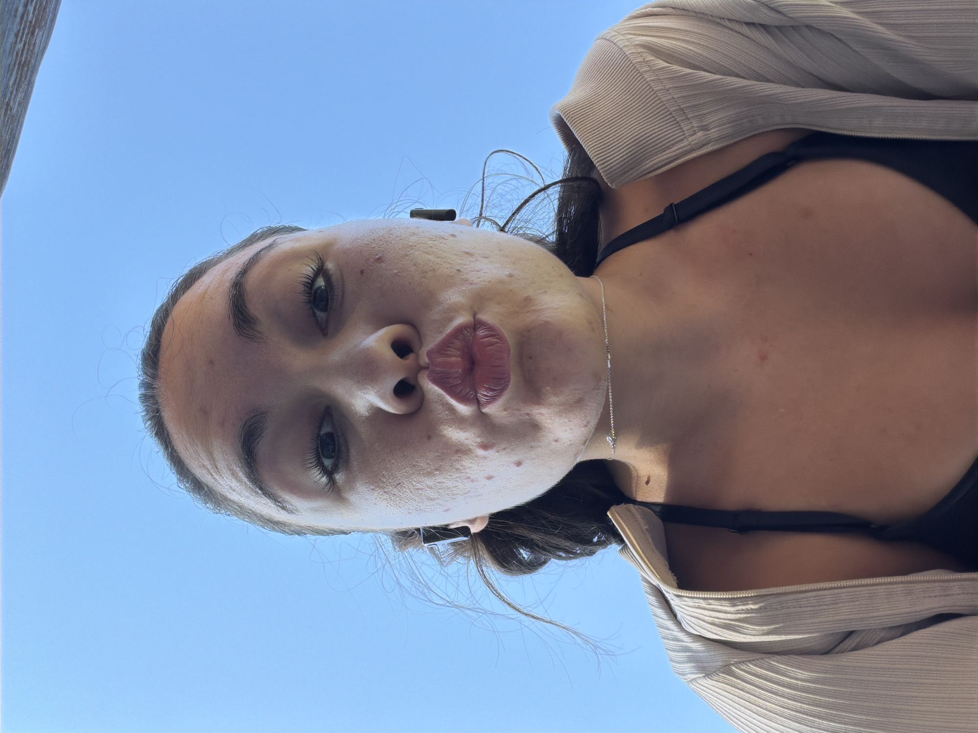 A low-angle, close-up, selfie-style shot shows a young woman against a clear blue sky. She is wearing a black bikini top and a beige, ribbed, zip-up cardigan. She has dark hair that is slightly windblown, and is wearing small black earbuds. Her lips are pursed for a kiss. The woman's face shows some acne. A thin silver necklace is visible around her neck. The image is taken outdoors, potentially on a balcony or rooftop, given the visible wooden railing at the top of the frame, suggesting an urban setting. The location is Varna, Bulgaria.