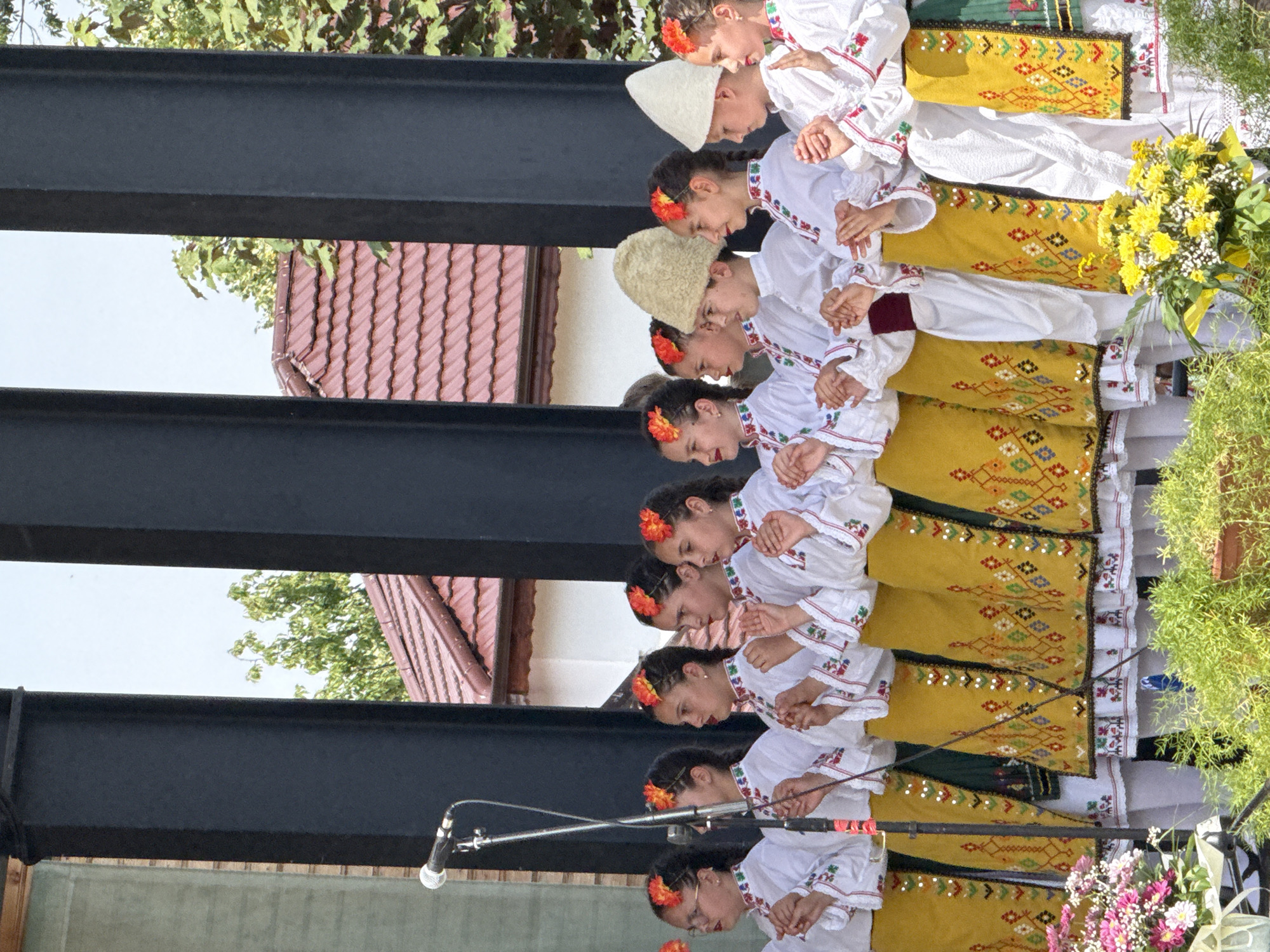 This is a medium shot photograph taken outdoors in Pordim, Bulgaria, capturing a group of young people performing on a stage. The performers are dressed in traditional Bulgarian folk costumes. There are at least eight individuals visible, all facing towards the right side of the frame and appearing to be singing or performing. They are arranged in two rows, with the front row of girls standing slightly in front of and between the individuals in the back row.
The costumes consist of white blouses with colorful embroidered trim around the collar, cuffs, and down the front opening. They are wearing brightly colored, patterned skirts or aprons over white skirts or dresses. Many of the girls have red and orange flowers in their braided hair. One individual in the back row is wearing a distinctive, tall, fuzzy hat, and another in the back row is wearing a conical white hat.
In the foreground, to the left, there is a microphone on a stand, suggesting a performance or presentation. There are also bouquets of yellow and pink flowers arranged at the base of the stage, adding to the festive atmosphere. Behind the performers, dark, vertical structural beams frame the scene. In the background, through the gaps in the structure, a residential building with a reddish-tiled roof and green trees are visible against a bright sky. The lighting suggests it is daytime and likely sunny.