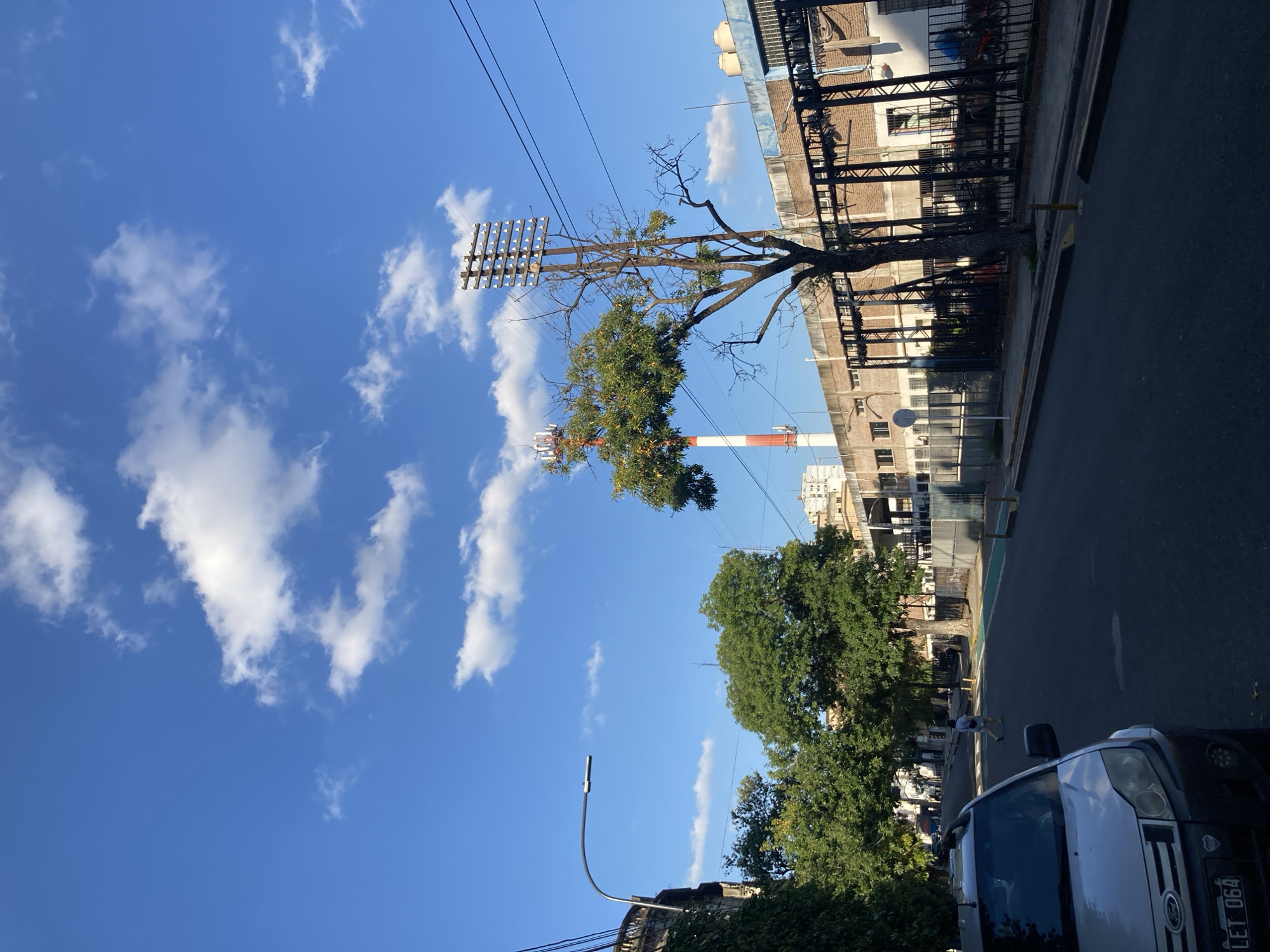 A street scene in Unknown, Unknown, featuring a light blue sky with puffy white clouds in the upper left. A tree in the mid-ground has a unique structure; it's partly bare with some green leaves. The tree supports a square grid of insulators. To the right of the tree is a brick building. Beyond the tree and building, a red and white chimney rises. The street occupies the right side of the frame and is paved with asphalt. A white vehicle with a license plate partially visible (LET 064) is parked at the bottom right. Further down the street, a person is visible in white clothing walking. Another tree with dense green foliage is in the mid-ground.