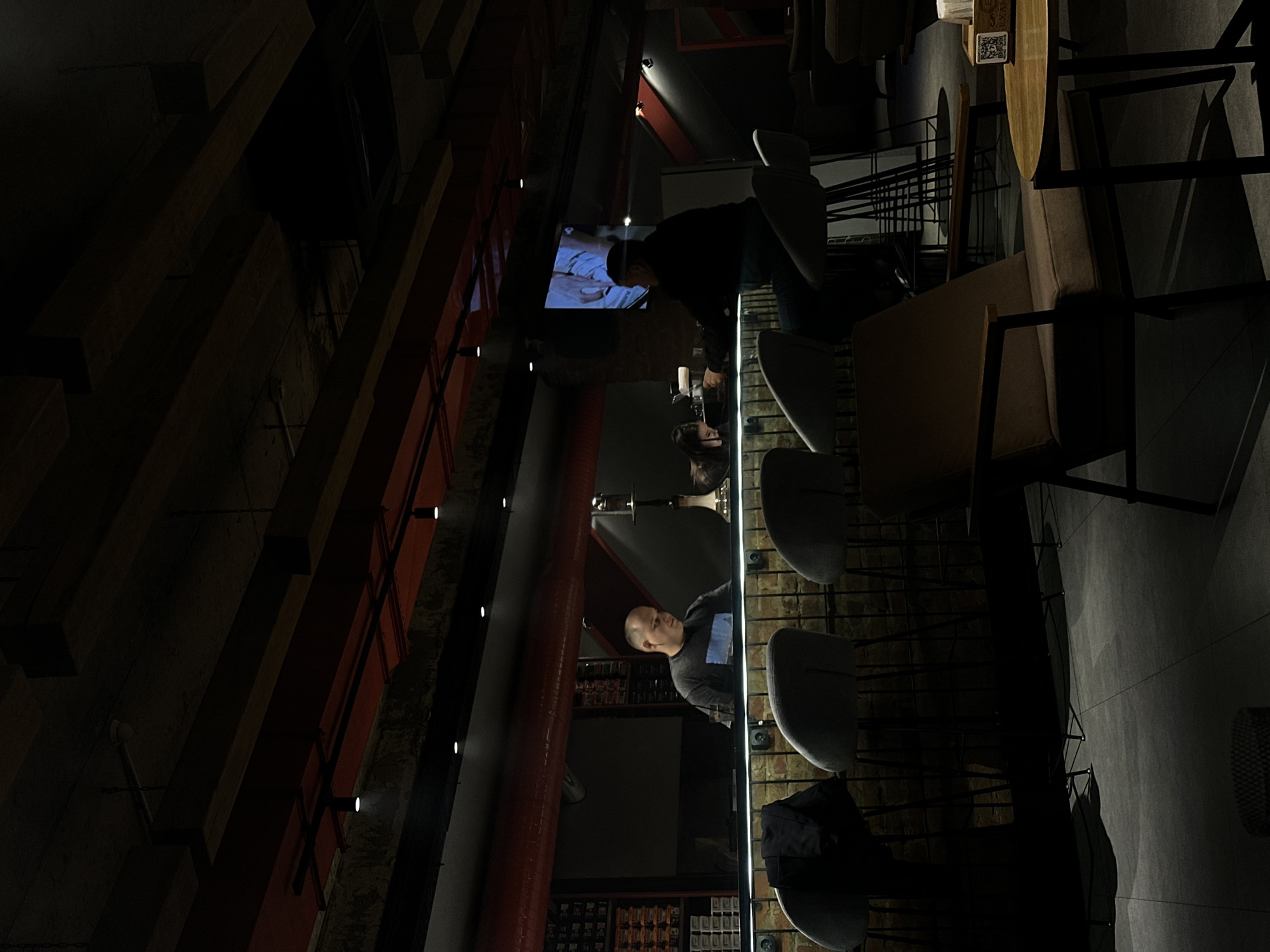 The image depicts the interior of a dimly lit bar or lounge located in Kazan, Russia. The establishment features an industrial-chic design with exposed dark wooden ceiling beams, red overhead ventilation pipes, and exposed brickwork behind the main bar counter. Numerous small, recessed spotlights provide illumination throughout the space.
In the mid-ground, a long, illuminated bar counter runs horizontally. Behind the bar, a bald male staff member, approximately 40-50 years old, wearing a dark t-shirt with a light graphic, stands looking down at the counter. To his right, partially obscured, a female with dark hair also looks down at the counter. A silver hookah with a hose is visible on the bar surface between them. Shelving units stocked with various items are visible on the wall behind the male staff member.
To the right of the frame, a male patron, approximately 30-40 years old, wearing a dark long-sleeved shirt, is seated on a light grey upholstered bar stool with a black metal frame, facing the left. Another female patron is partially visible seated further left at the bar. Several identical bar stools are positioned along the counter, with a black jacket draped over one on the far left.
Above the right section of the bar, a flat-screen television is mounted on the wall, displaying an image that appears to be a person's hands.
The flooring consists of dark grey tiles. In the foreground to the right, a lounge area is visible with a brown upholstered armchair featuring wooden armrests. Adjacent to the armchair is a small, round wooden table with a black metal base. On the table, there is a small wooden box containing tissues or napkins, displaying a QR code. Shadows are cast across the floor from the furniture and bar stools, indicative of the directional lighting. A smoke detector is visible on the ceiling in the upper left quadrant of the image.