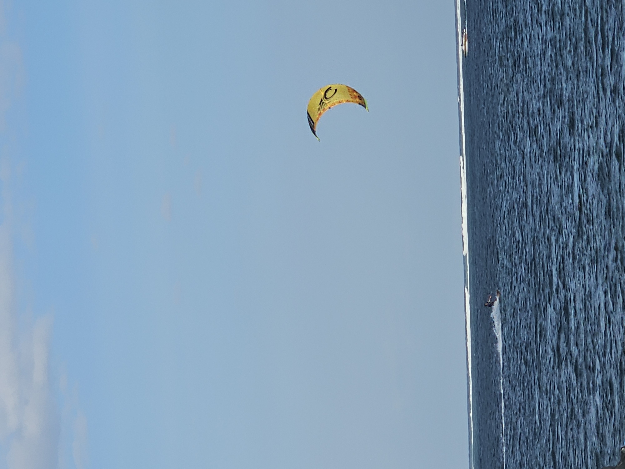 A kite surfer is visible on the choppy, dark blue waters of the ocean, leaving a white spray trail as they move from left to right. A yellow and orange kite is suspended in the light blue sky above and to the right of the kite surfer. In the far distance, a white line of breaking waves marks the horizon, and a small boat with an orange stripe is visible to the right of the kite surfer. The scene appears to be a coastal area in Denpasar, Indonesia.