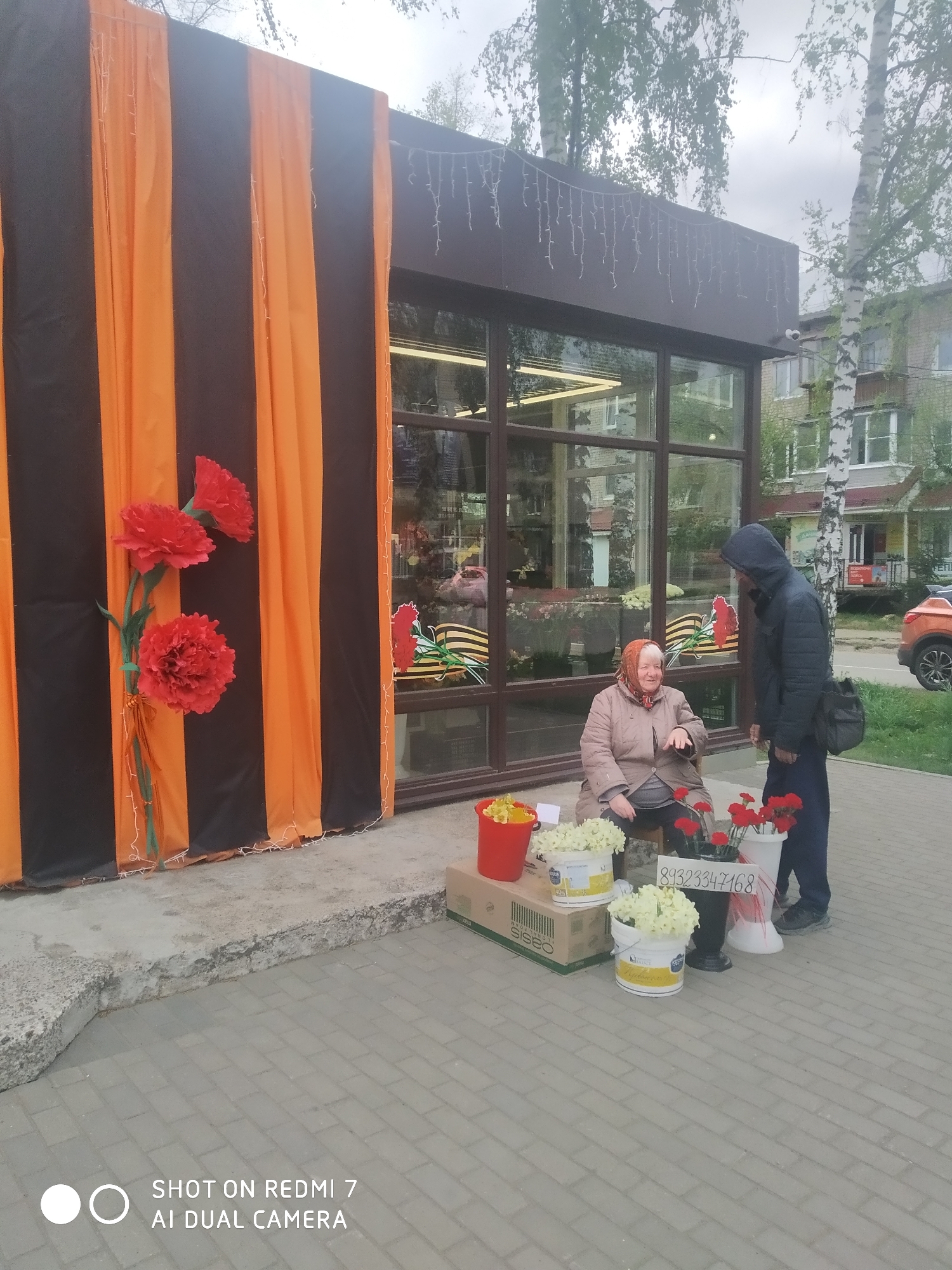 Outside a building with large glass windows, a woman sits on a stool selling flowers. She is wearing a light brown jacket and a headscarf. In front of her, there are buckets of white and red flowers, along with a sign displaying the phone number "89323347168". A man wearing a dark jacket with a hood up and a shoulder bag is standing in front of her, seemingly interacting with her. The building is adorned with orange and black striped decorations and large red paper flowers. A St. George ribbon is also visible in the window. The location is Chaikovsky, Russia, as indicated by the phone number's regional code. The photo was taken with a Redmi 7 AI Dual Camera.