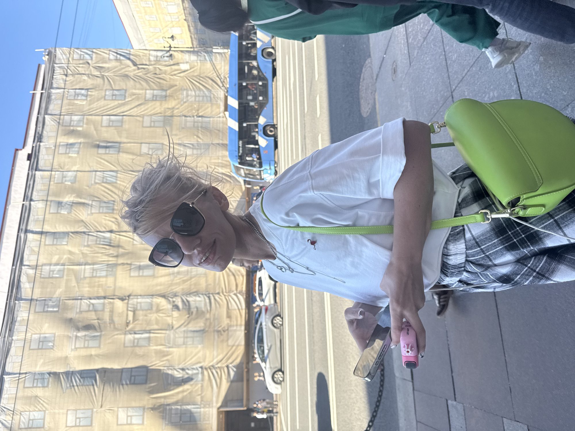 A blonde woman with sunglasses and a white t-shirt stands on a sidewalk in Saint Petersburg, Russia, holding a phone and a pink object. She is wearing checkered pants and a green purse. In the background, a blue trolleybus is on the street, and a building covered in scaffolding is visible. The sky is clear and blue.