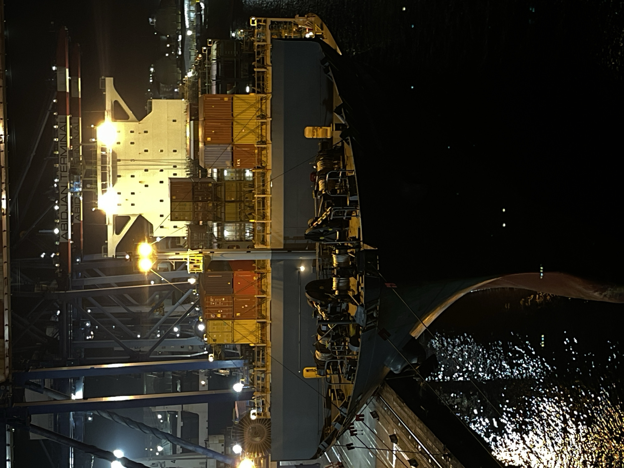 The image captures a large cargo ship docked at the Abidjan Port, Ivory Coast, at night. The ship is laden with numerous shipping containers, stacked high on its deck. The port infrastructure, including cranes and terminal buildings, is visible in the background, illuminated by artificial lights. The water surrounding the ship reflects the lights, creating a shimmering effect. The overall scene depicts a busy, operational port at night, with the cargo ship appearing to be in the process of loading or unloading.