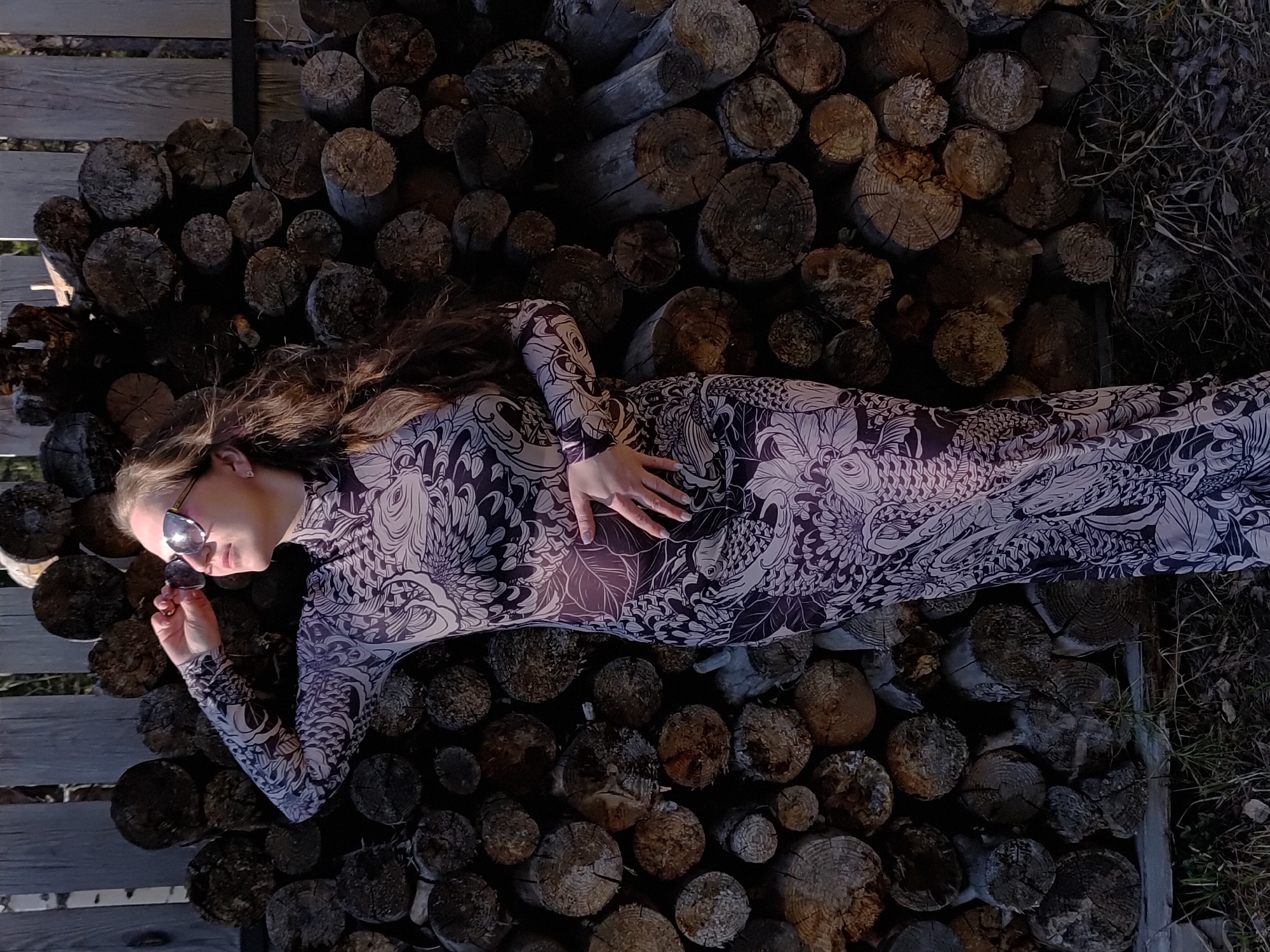 A young woman wearing a long-sleeved dress with a black and white floral pattern and sunglasses is lying on a pile of firewood. The firewood consists of various sized logs, neatly stacked. She is holding her sunglasses in her left hand. The location appears to be outdoors, near wooden planks and sparse vegetation. The specific city is unknown, despite its mention in the prompt.