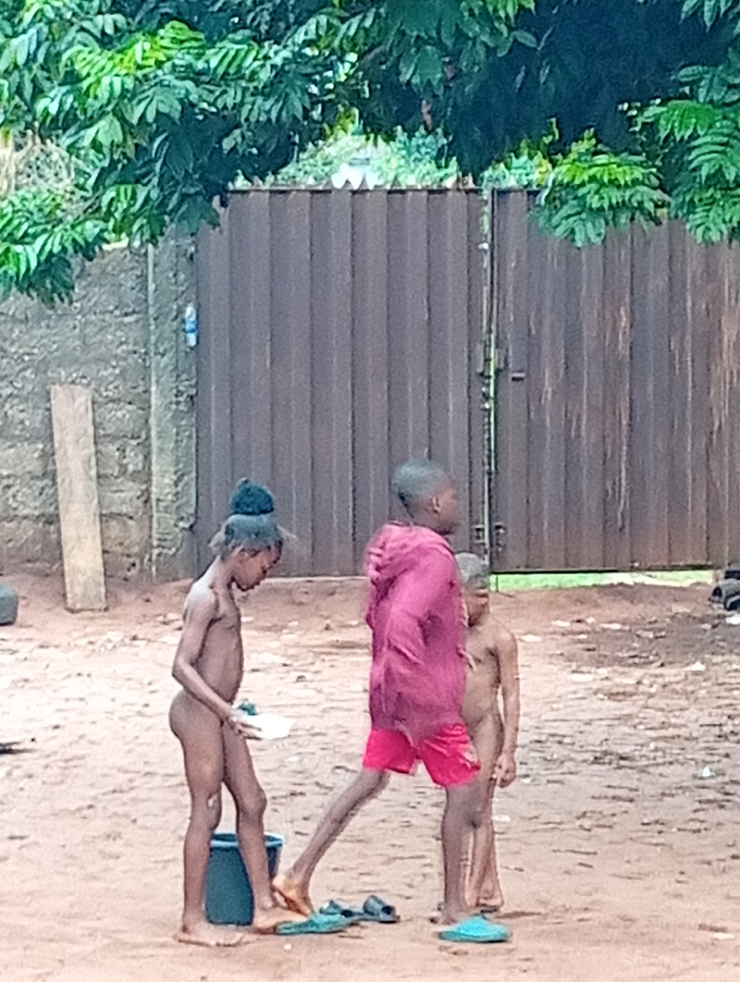 The image depicts three children outdoors. Two are nude and one wears a red hooded jacket and red shorts. The nude children stand near a blue bucket and some sandals. One child is holding a white object. The environment appears to be a sandy or dirt-covered area with a brick wall and a corrugated metal fence in the background. Green foliage is visible overhead. The location is described as Ogidi, Nigeria.