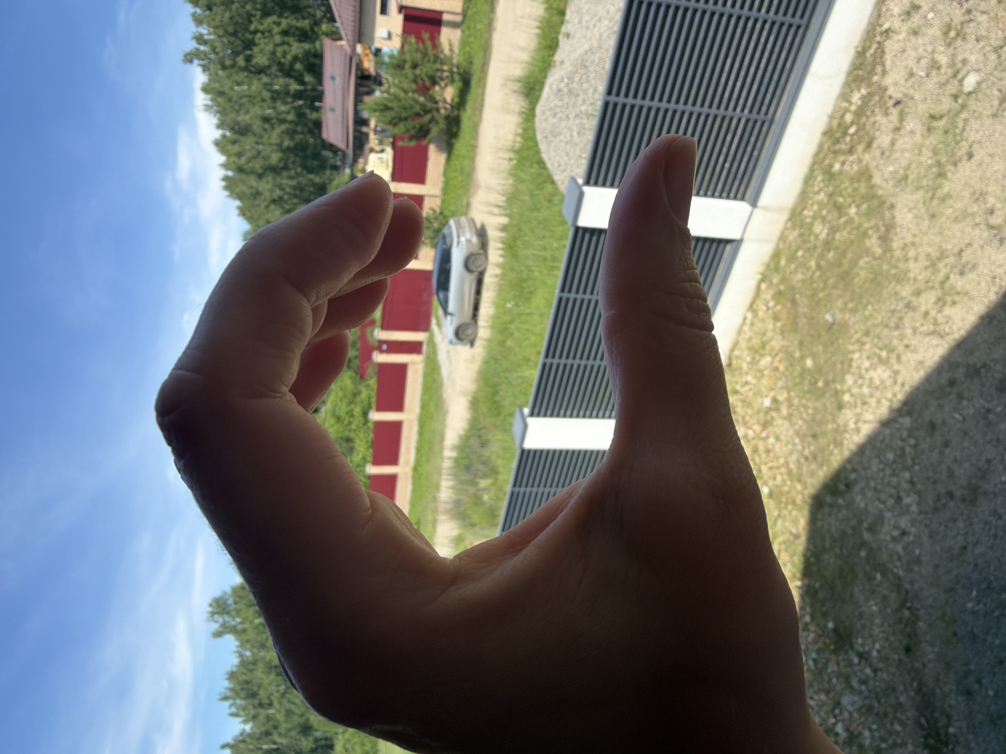 The image shows a close-up of a person's hand, with fingers curled to form a C-shape, obscuring much of the background. The background depicts a rural or suburban scene with a clear blue sky dotted with wispy clouds. Green trees line the horizon, behind which several houses with brown roofs are visible. A light-colored car is parked on a dirt road in the middle ground. In the foreground, a grey, slatted fence runs horizontally across the frame. The lighting suggests it is daytime, likely sunny, casting shadows on the ground. The specific location is identified as Malinovka, Russia.