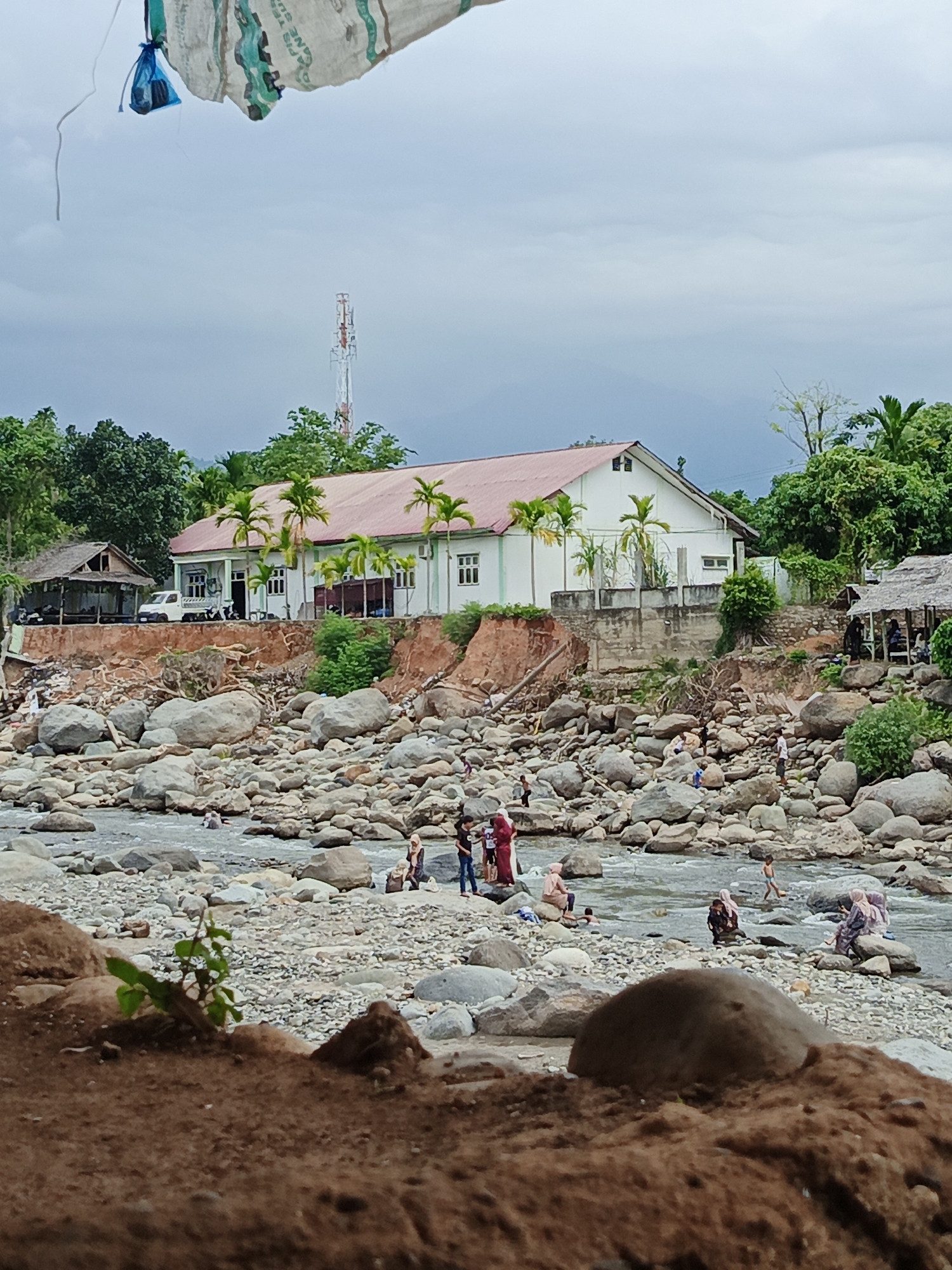 This detailed image captures a daytime scene at a rocky riverbank in Meurah, Indonesia, under an overcast sky. In the immediate foreground, dark brown soil and a small green plant are visible, with a large, rounded dark rock partially obscured. The midground features a wide, shallow riverbed densely covered with numerous grey boulders and smaller rocks, through which water flows. Several individuals are present in and around the river; some are wading in the water, others sit on the rocks. People are dressed in casual attire, with several women wearing hijabs.
On the riverbank, an eroded dirt slope transitions to a more elevated area. To the left, a simple, open-sided wooden structure with a corrugated roof stands next to a white pickup truck. Behind this, a prominent white building with a red-orange gabled roof is situated on a raised, concrete foundation. Palm trees are planted in front of this building, which features multiple windows and a porch area. The bank below the building shows significant erosion, exposing reddish soil and scattered debris. To the right of the white building, another smaller, thatched-roof structure is visible, with people gathered inside or nearby.
Beyond the buildings and structures, dense green foliage and trees line the horizon, with mountains faintly visible in the far distance. A tall, white communication tower with multiple antennas rises above the treeline on the left side of the background. At the very top edge of the image, partially obscured, is a piece of light-colored fabric or tarp with green markings, from which a small blue plastic bag hangs.