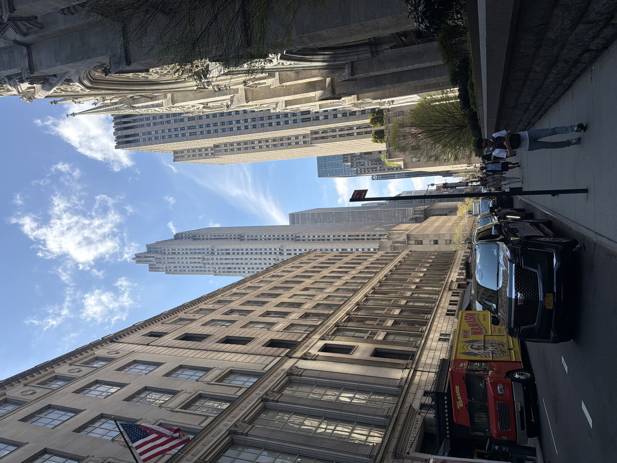 The image is taken on a street in New York City, United States of America. The camera faces upward, capturing tall buildings and a patch of sky with light clouds.
On the left side of the street is a stone building with multiple windows and an American flag hanging. Adjacent to it is a food truck and a black SUV parked on the street.
On the sidewalk, there are several pedestrians walking. The sidewalk is lined with a stone wall.
On the opposite side of the street, there is a cathedral-like structure with intricate stonework. Further down the street, several high-rise buildings are visible. The sky is blue with wispy clouds.