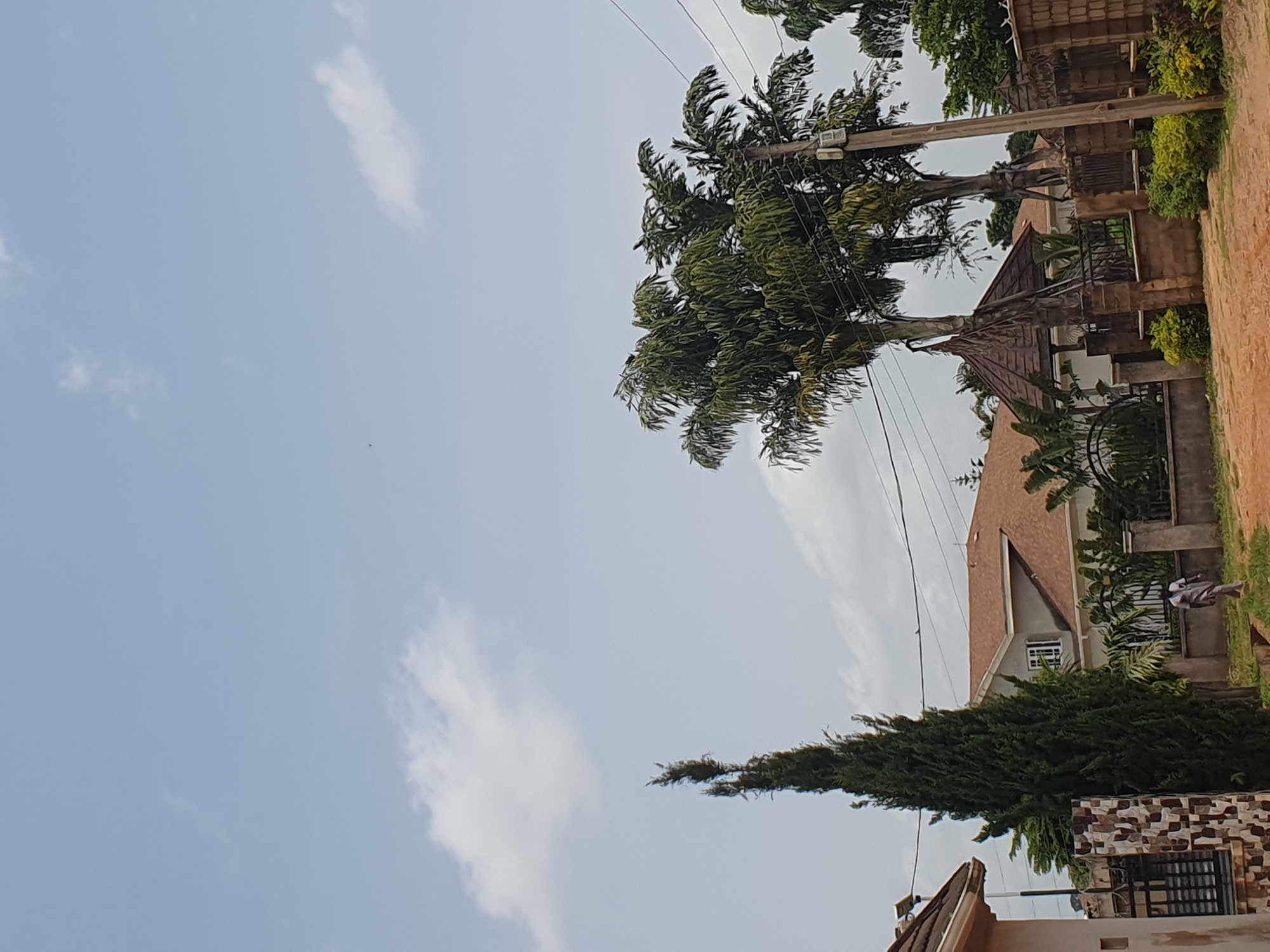 The image depicts a residential street scene in Gura Top, Nigeria, under a pale blue sky with scattered white clouds. In the foreground, a dirt road curves past a low wall topped with decorative iron fencing. Beyond the wall, a man in a light grey traditional outfit walks away from the camera. To the left, a tall, slender cypress tree stands prominently, adjacent to a house with a patterned stone facade and dark metal fencing. In the background, a large house with a brown, tiled roof is visible, surrounded by lush green vegetation and palm trees. A hexagonal wooden gazebo with a pointed roof is situated in front of the house. Utility poles with overhead wires are also present in the scene, indicating the presence of electrical infrastructure. The overall impression is of a quiet, suburban environment with tropical flora.