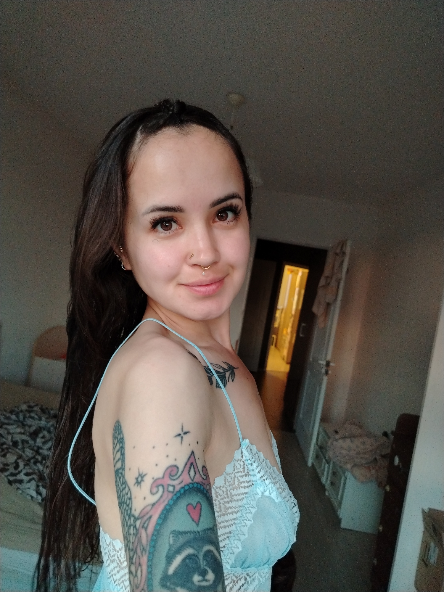 A young woman with long, dark hair and brown eyes is shown in a medium close-up shot, looking towards the camera. She is wearing a light blue lace bralette. She has piercings on her septum and right nostril. She has tattoos on her left arm, including an image of a raccoon within a decorative frame and a sprig of leaves on her shoulder. The background appears to be a bedroom, with a bed visible on the left and a doorway leading to another room on the right. The location is identified as Noviy, Russia.