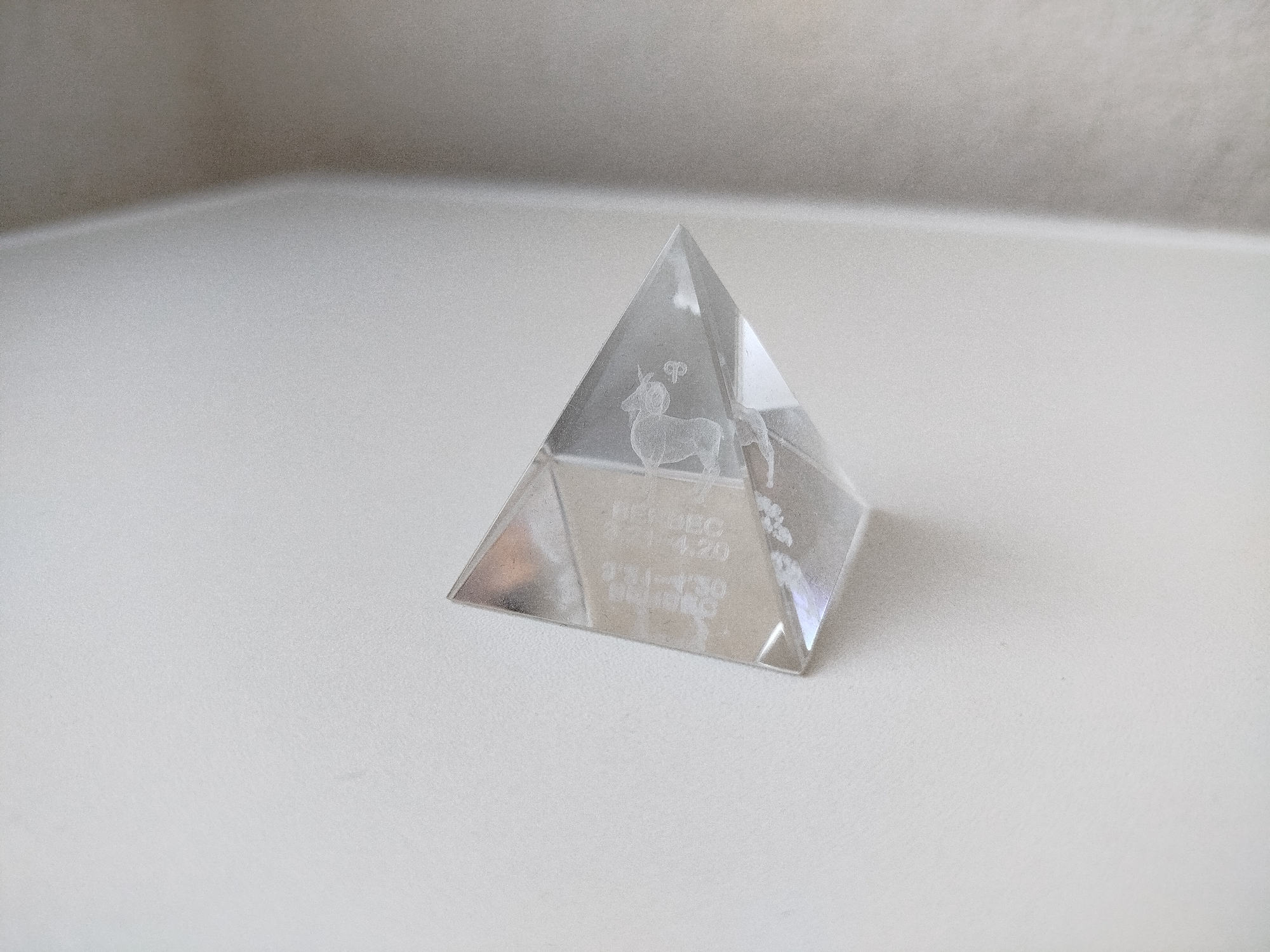 A clear, triangular pyramid object rests on a white, flat surface. The pyramid features a depiction of a goat, likely representing the astrological sign Capricorn, and dates "Dec 21-Jan 20". The pyramid also contains faint floral-like elements, possibly decorative. The background is out of focus, showing a light-colored wall. The location cannot be inferred from this image.