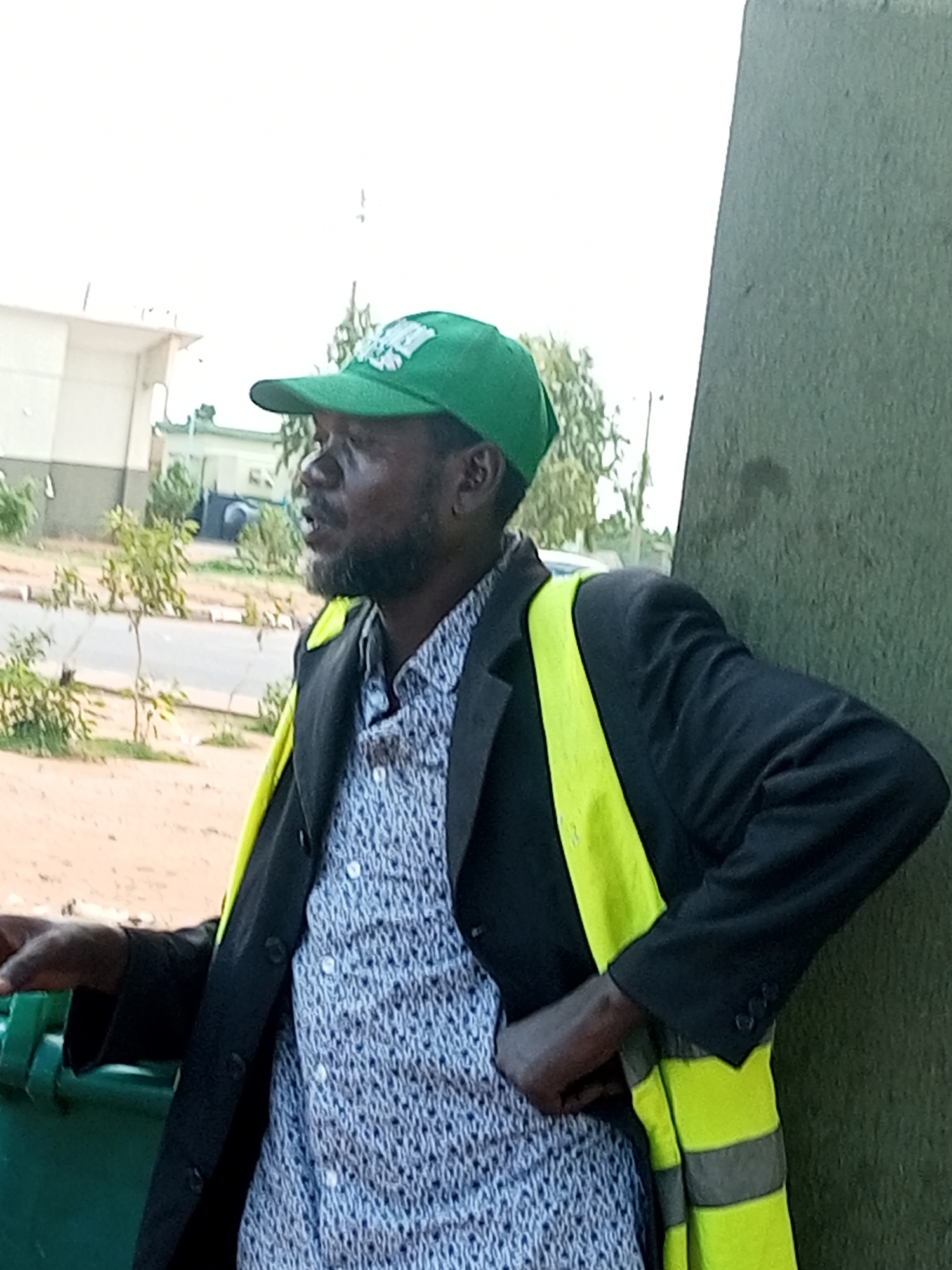 A man wearing a green cap with white lettering and a black jacket over a patterned shirt is standing in front of a green wall in Gombe, Nigeria. A high-visibility yellow vest with reflective stripes is worn over his jacket. His left arm is bent at the elbow, and his hand is resting on a green bin. In the background, there are some trees, a road, and buildings. The man has a beard and is looking to his right with his mouth slightly open. The lighting suggests it is daytime.