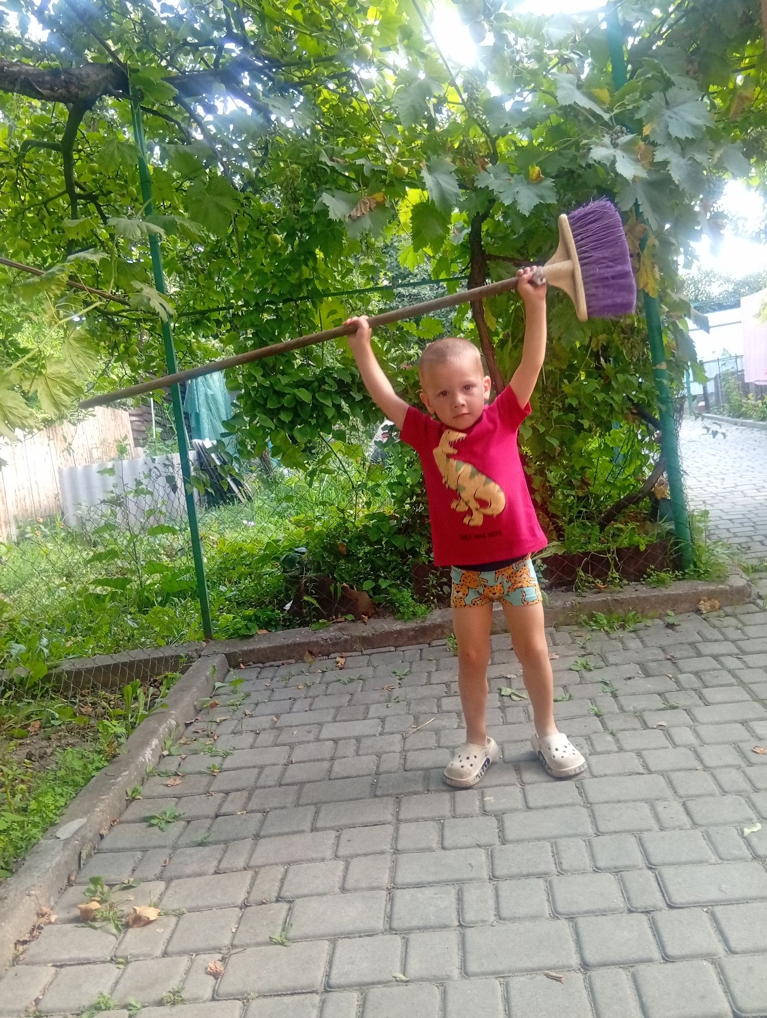 A young boy, appearing to be around four years old, stands outdoors on a paved area, holding a broom horizontally above his head with both hands. He is wearing a red t-shirt with a yellow dinosaur graphic and patterned shorts. The boy is barefoot, wearing beige Crocs-style shoes. The background consists of lush green foliage, a wooden fence, and a chain-link fence. The paved surface is made of interlocking rectangular stones, with some scattered leaves and grass growing between them. The overall scene suggests an outdoor play or activity in a residential or garden area.