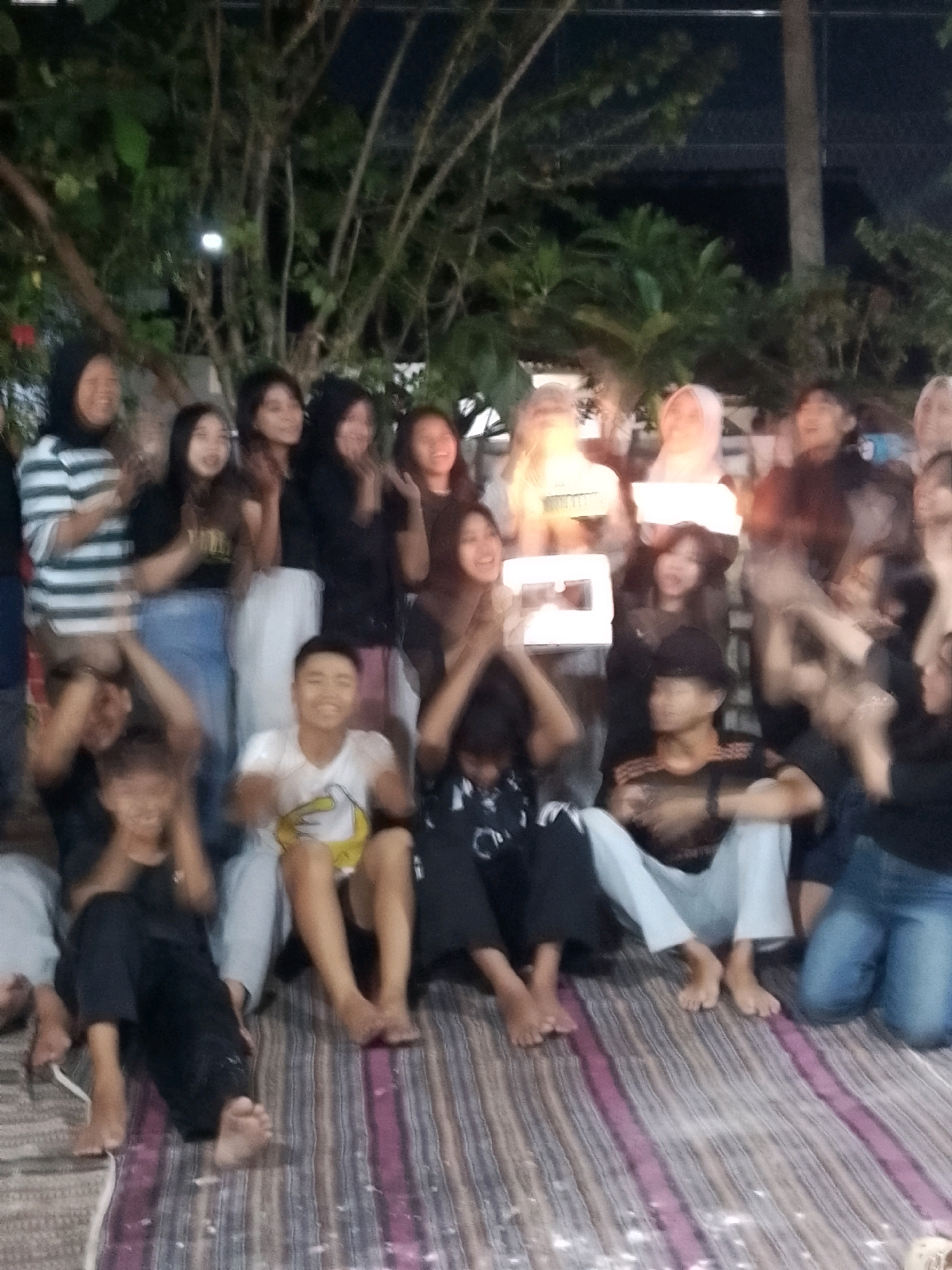 The image captures a group of at least twelve young people, appearing to be teenagers or young adults, gathered outdoors at night. They are seated and standing on what appears to be a patterned mat or rug, likely in a residential area or a casual outdoor gathering space. The lighting is dim, suggesting it is evening or nighttime, with some artificial light sources illuminating the scene.
Several individuals are holding up illuminated objects, possibly phones or small lights, creating a glowing effect. The people are dressed in casual attire, including t-shirts, long-sleeved shirts, and jeans. Some are wearing headscarves. The background consists of dark foliage and trees, with a visible pole that could be a street light or part of a building structure. The overall atmosphere appears to be one of social gathering and shared activity. The blurry nature of the image indicates it might have been taken with a low-quality camera or in low light conditions, or that the subjects were in motion. There is no explicit indication of the city of Jember, Indonesia, in the image itself, but this context is provided externally.