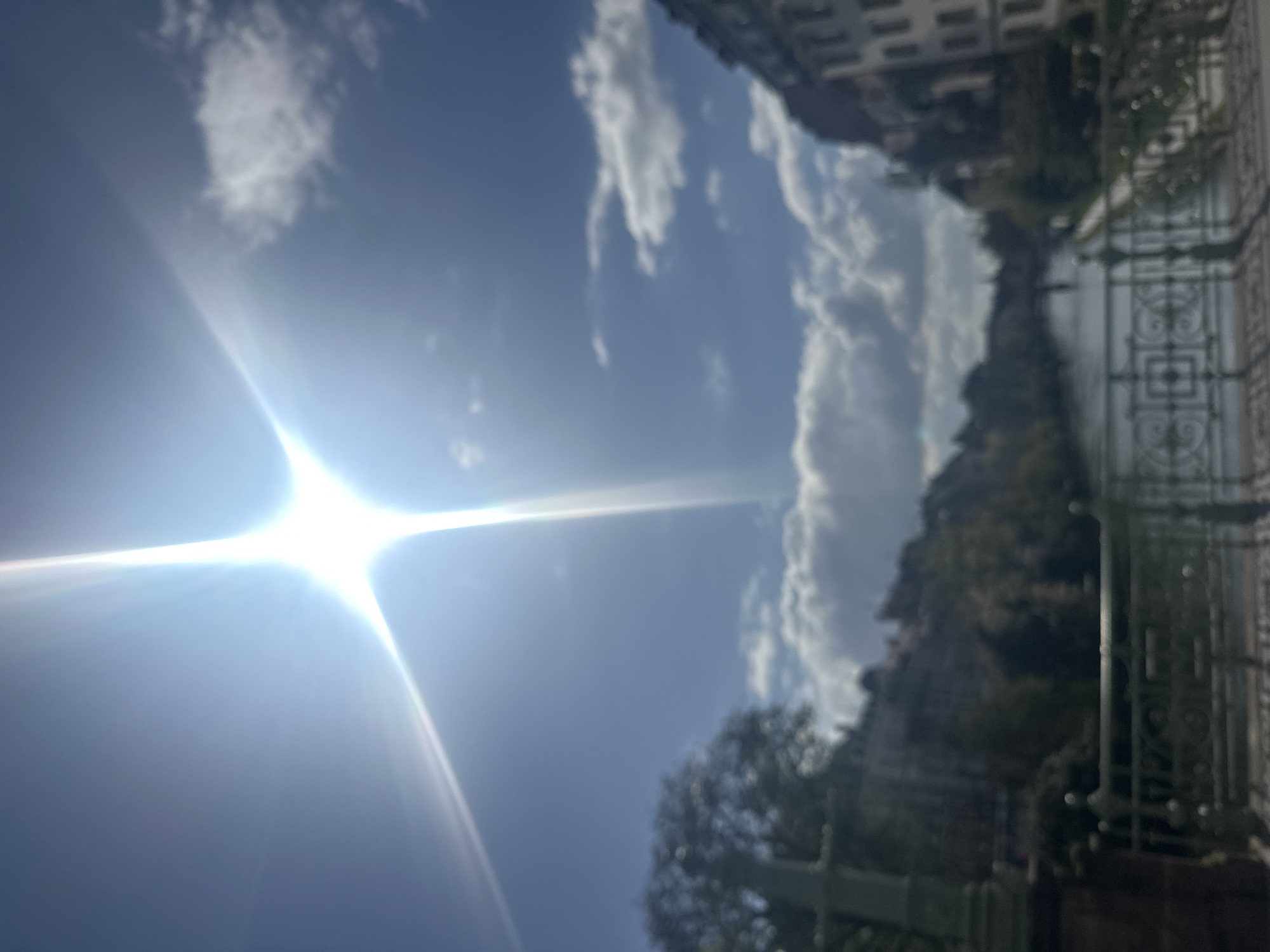 The image captures a cityscape in Strasbourg, France, along the Quai des Bateliers. The sky is predominantly blue with scattered clouds, and the sun is shining brightly, creating lens flare. Adjacent to the Quai des Bateliers is the canal, with greenery and trees lining the banks. A decorative wrought-iron fence runs along the quayside. Buildings are visible in the background, including typical Strasbourg architecture. The scene is taken from a low angle, looking up.