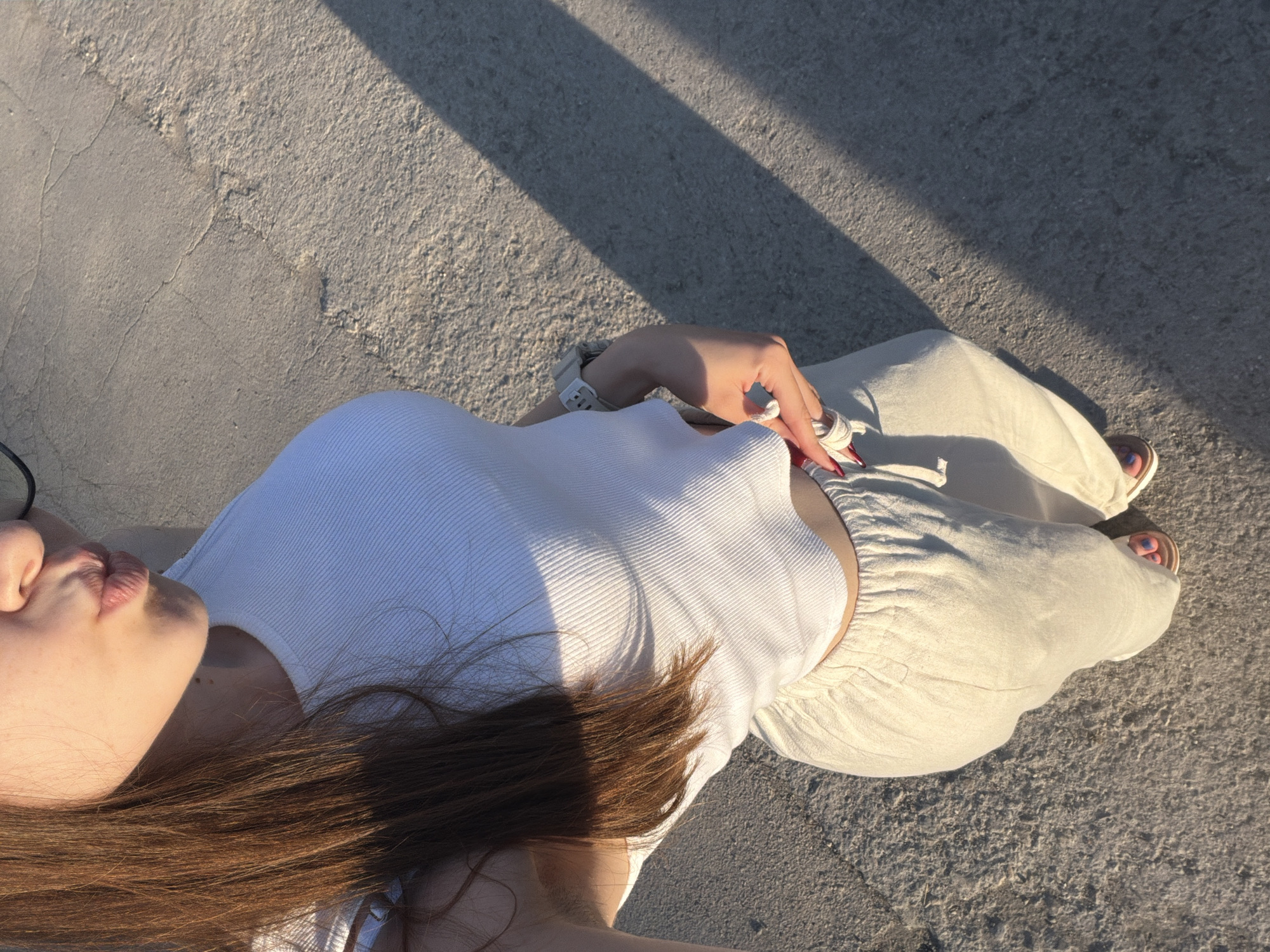 A young woman with long, brown hair and fair skin is standing outdoors, facing the camera with her lips pursed in a kissing gesture. She is wearing a white ribbed crop top and loose-fitting, light-colored pants. She has a watch on her left wrist and is holding the waistband of her pants with her right hand. She is wearing sandals on her feet. The ground beneath her appears to be paved with concrete. A distinct shadow from an unseen object is cast across the ground to her right. The lighting suggests it is either sunrise or sunset. The setting is likely urban, given the paved surface, and could be a sidewalk or plaza. The style of the paving stones is consistent with those found in public spaces in the city of Varna, Bulgaria.