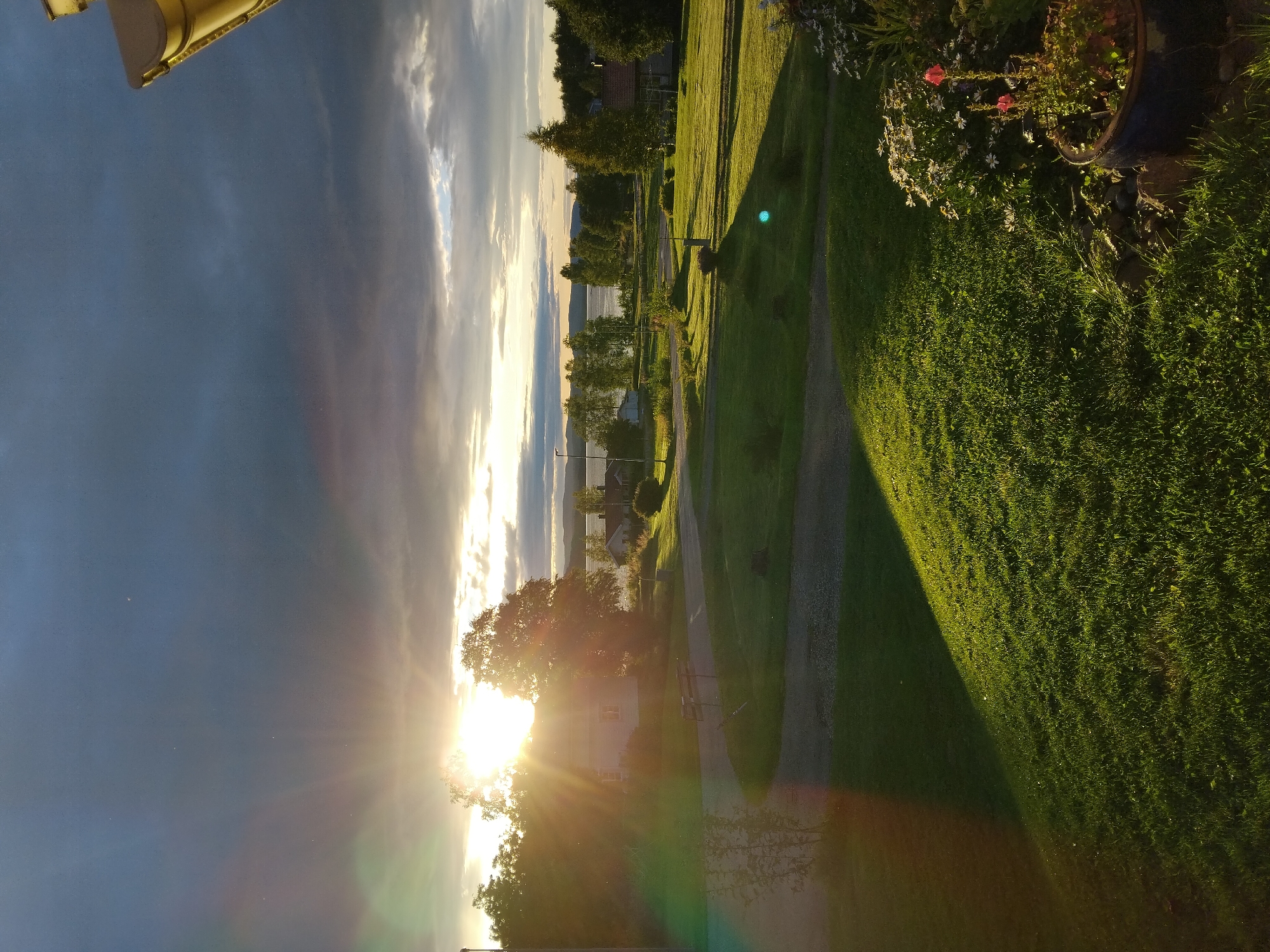 The image captures a serene landscape scene at sunset in Rätan, Sweden. The sun is low on the horizon, casting a warm glow and creating lens flare effects. Dark, dramatic clouds fill the upper portion of the sky, contrasting with the bright, warm light of the sunset peeking through. In the middle ground, a body of water, likely a lake, is visible, with a treeline and a few houses scattered along its edge. The foreground is dominated by a lush green lawn, with a paved path curving through it. A large tree stands prominently in the middle ground, partially obscuring a white house. The overall impression is one of a peaceful, rural evening.