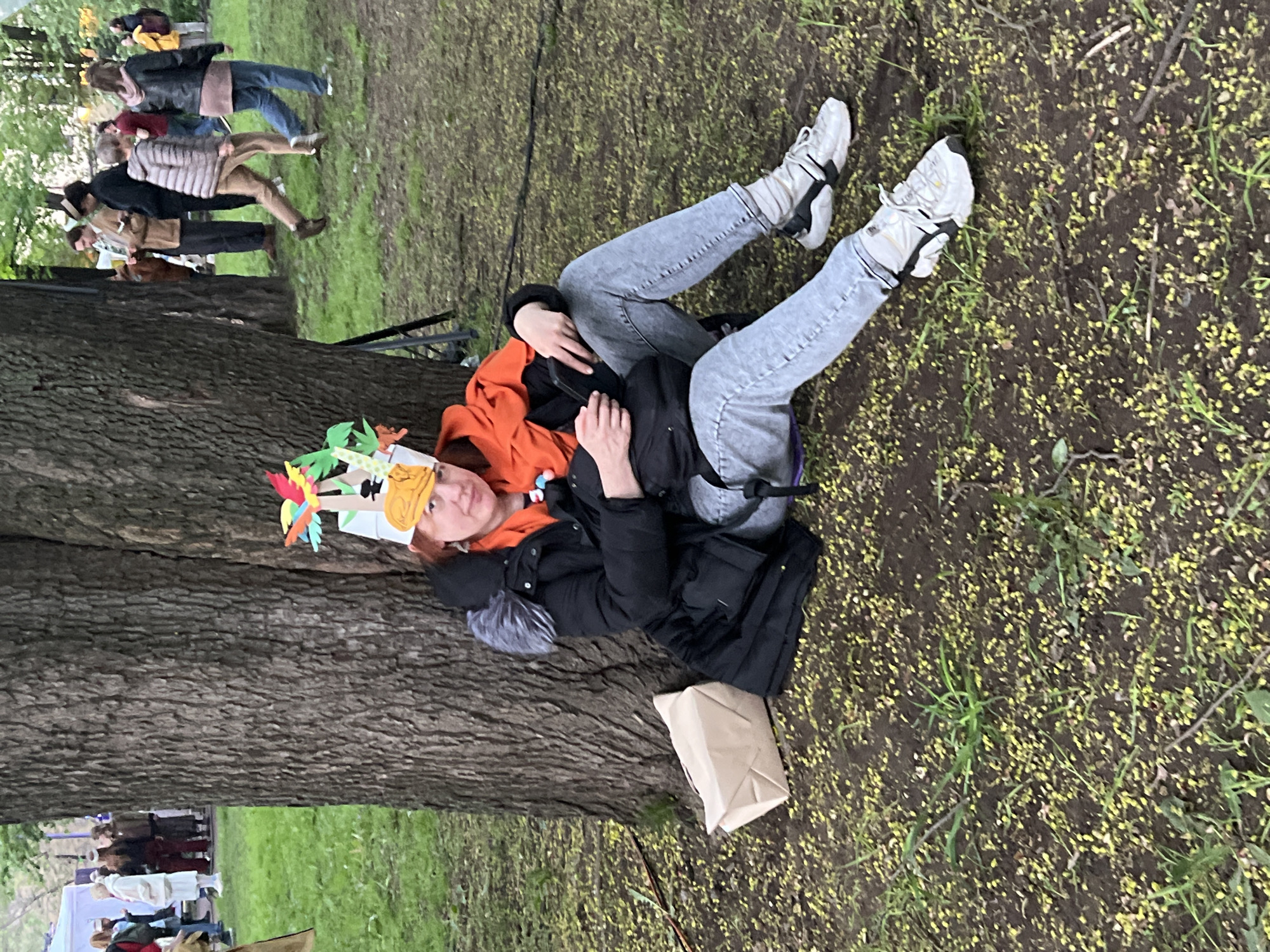 A person lies prone near the base of a large tree in what appears to be a park or grassy area. The individual is wearing light gray jeans, white sneakers with black accents, an orange top, and a black jacket with a fur-trimmed hood. They also sport a whimsical paper crown with colorful cut-out shapes. A brown paper bag rests near the tree trunk next to the person.
The ground is covered in dirt, grass, and small yellow flower petals or pollen. In the background, several people are walking or standing in a more distant grassy area.
Based on the style of clothing, urban park setting, and presence of small yellow flowers or pollen which are common in spring, it's possible this photograph was taken in a park in Saint Petersburg, Russia. The trees and foliage are typical of a park in that region. It's important to note that this location is speculative.
