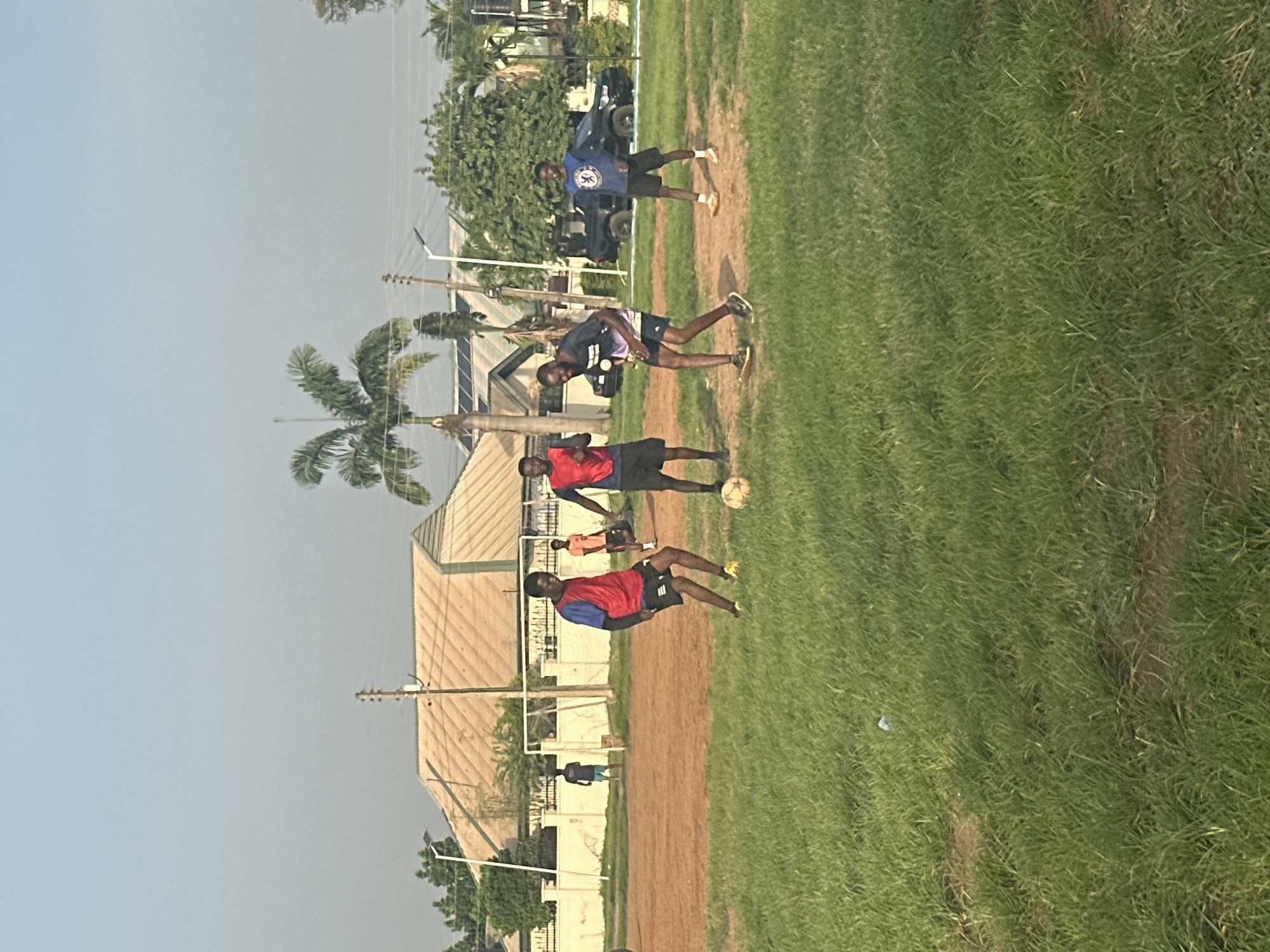 This media file captures a group of young men engaged in a soccer practice on a grassy field in Abuja, Nigeria. The scene appears to be set in the late afternoon, as indicated by the soft, warm sunlight casting long shadows and the clear, pale blue sky. The weather is pleasant, with no visible clouds.
The primary subjects are the four individuals, all appearing to be of African descent, dressed in athletic attire. They are spread across the field, some actively participating in the game and others observing. One person is in the foreground, wearing a red and blue jersey and black shorts, with a soccer ball at his feet. Another individual, in a dark blue and purple jersey with black shorts, is positioned behind him, seemingly about to interact with the ball. Further back, two other men are visible. One wears a blue sweatshirt with a crest and black shorts, while the other is dressed in an orange t-shirt and black shorts. The activity is clearly a soccer training session, with the players demonstrating focus and athleticism.
The setting includes a well-maintained grassy field, likely part of a sports facility, with a dirt track bordering parts of it. In the background, several buildings with distinctive, light-colored, angled roofing are visible, suggesting a residential or institutional area. A prominent palm tree stands to the left, reinforcing the tropical or subtropical environment of Nigeria. Utility poles and wires are also present, indicating the presence of infrastructure. A dark-colored SUV is parked in the background, adding to the everyday scene. The overall mood is one of energetic activity and community engagement, characteristic of a sports practice. No visible text is present in the image.