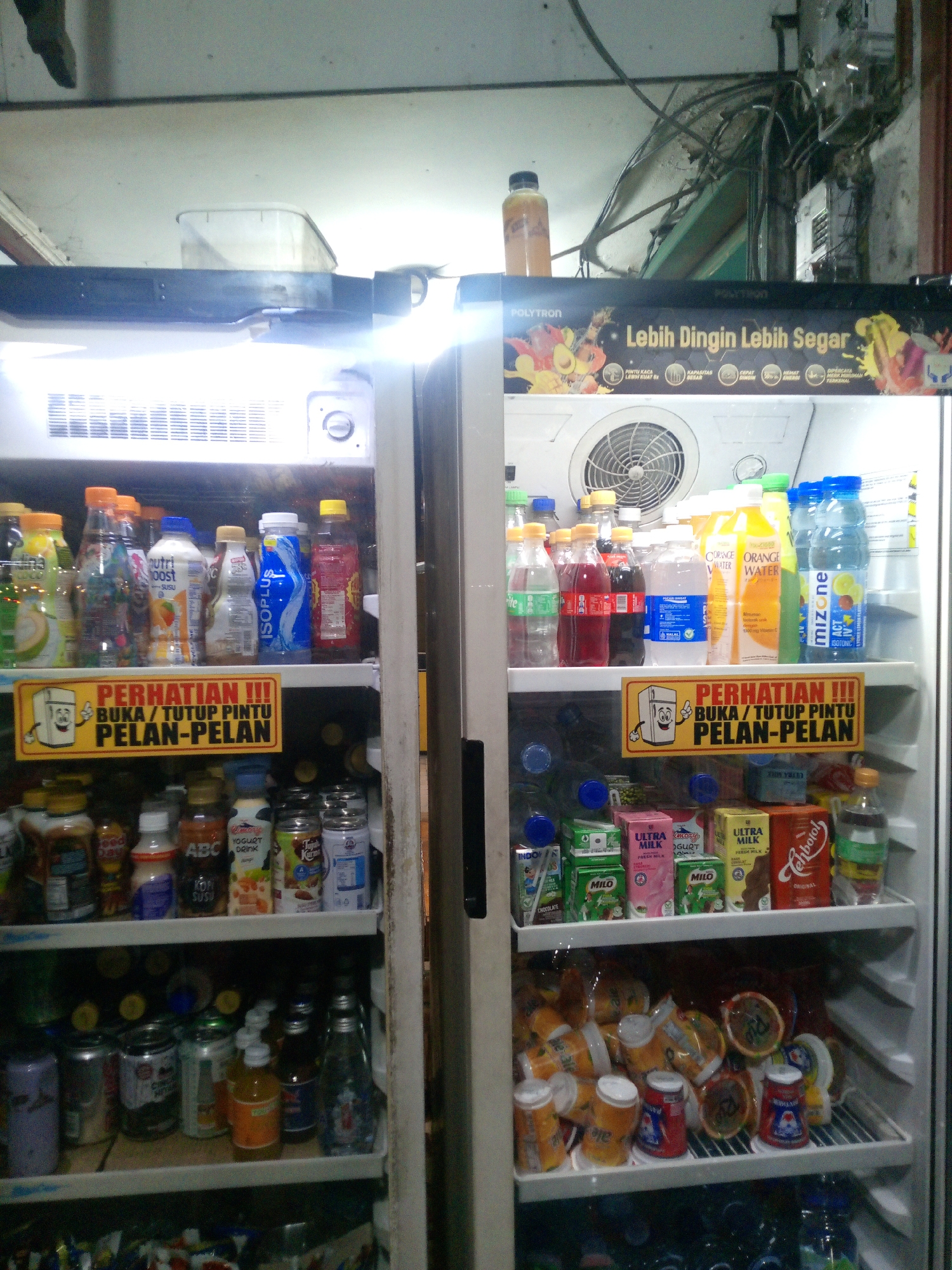 The image depicts an open refrigerator in what appears to be a small convenience store or warung in Bitung Jaya, Indonesia. The refrigerator is well-stocked with a variety of beverages, including bottled water, soft drinks, and juice boxes. Several shelves are visible, filled with products arranged in a somewhat organized manner. On the top shelf of the left side, a plastic container is visible. Above the shelves, a banner reads "POLYTRON" and "Lebih Dingin Lebih Segar" (Colder, Fresher). A sign with an illustration of a refrigerator and text that reads "PERHATIAN !!! BUKA / TUTUP PINTU PELAN-PELAN" (Attention!!! Open / Close Door Slowly) is prominently displayed on the middle shelf of the left side. The right side of the refrigerator shows more drinks and some stacked boxes on the bottom shelf. The overall environment suggests a retail setting where drinks are kept chilled for sale. There are no people visible in the image.
