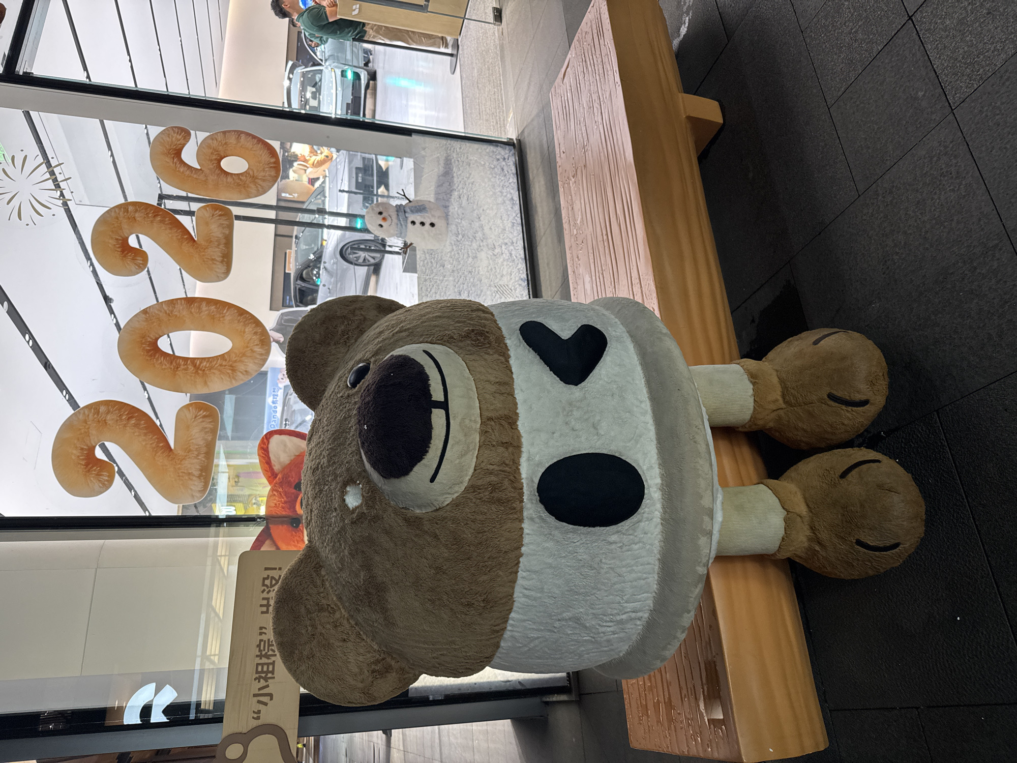 The image captures a scene in Jinjiang District, China, featuring a large, plush koala bear resting on a wooden bench. The koala is light brown, with a white belly marked with a black heart and a black circle. The bear's face is predominantly brown with black features. In the background, visible through a window, are the numbers "2026" in large, fluffy digits. The window also reflects a snowman figure and a partial view of what appears to be a commercial interior, possibly a mall or store, with people visible in the upper portion of the reflection. A vertical sign with Chinese characters is positioned near the window. The floor is covered with dark gray tiles. The bench supporting the koala is made of light brown wood and has a simple, slatted design. The image is taken from a slightly elevated perspective.