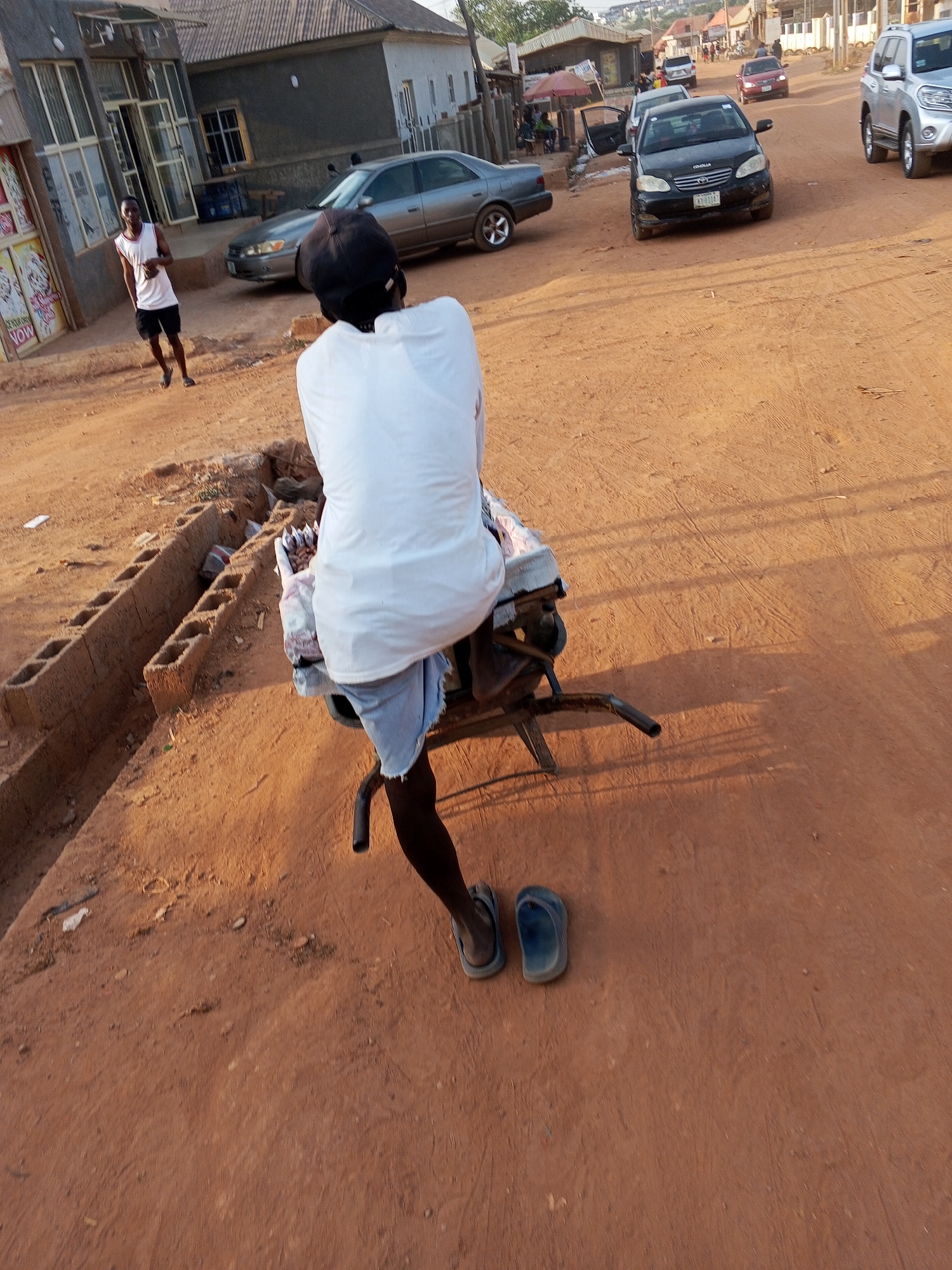 The photo shows a person sitting on a wheelbarrow on a dirt road in Abuja, Nigeria. The person is wearing a white shirt, shorts, and sandals, with a cap. Behind the person is a building with a store in front of it, and a person wearing a white tank top and black shorts is standing in front of the store. In the background, there are several cars parked along the street. The road is red dirt.