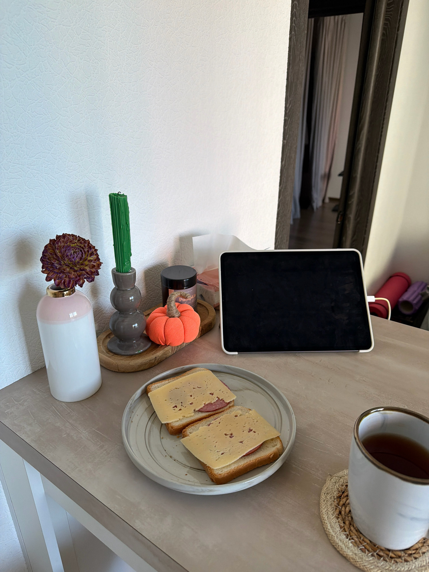The image depicts a close-up, indoor scene on a table. In the foreground, a white mug with a gold rim rests on a woven coaster. To the left of the mug, a gray plate holds two open-faced sandwiches with slices of cheese and what appears to be cured meat. Further to the left, a vase with a pink band at the top contains a dark purple, dried flower. Adjacent to the vase is a gray candlestick holding a tall, green, ribbed candle. In front of the candlestick sits a small, bright orange, fabric pumpkin with a twisted stem. Behind these items, a black tablet or e-reader is propped up. The background shows a textured white wall and a doorway with brown trim, revealing a glimpse of curtains and possibly exercise equipment in an adjacent room. The overall lighting suggests a daytime setting. The location is described as Kyiv, Ukraine.