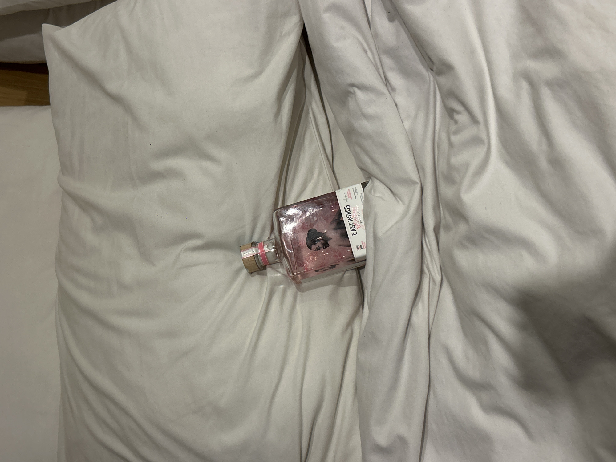 A close-up, high-angle shot depicts a bed with white sheets in disarray. A partially obscured square bottle of East Indies gin is resting on the left side of the bed. The location is not discernible in the image. The specific type of bed or the room it is in cannot be determined from this view. There are no people or actions visible.