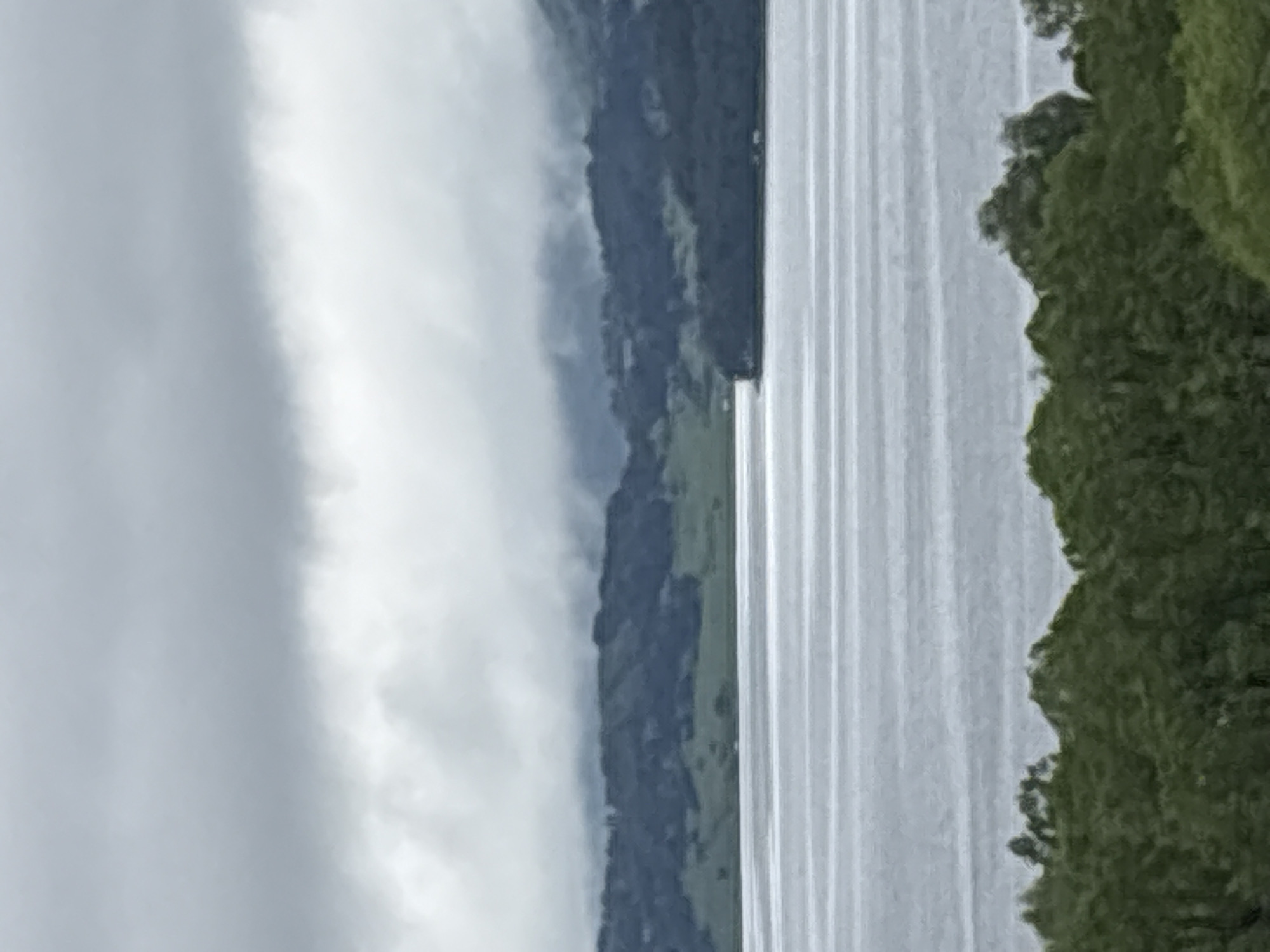 The image shows a body of water, likely a lake or reservoir, with repeating vertical lines on the surface suggesting ripples or small waves. On the right side of the water, there's dense green vegetation, likely trees. On the left, there is a hillside or mountain partially obscured by heavy clouds or fog. The presence of mountains and a large body of water suggests a lakeside or mountainous environment. There is no evidence to confirm the location is in Tsigov chark, Bulgaria.