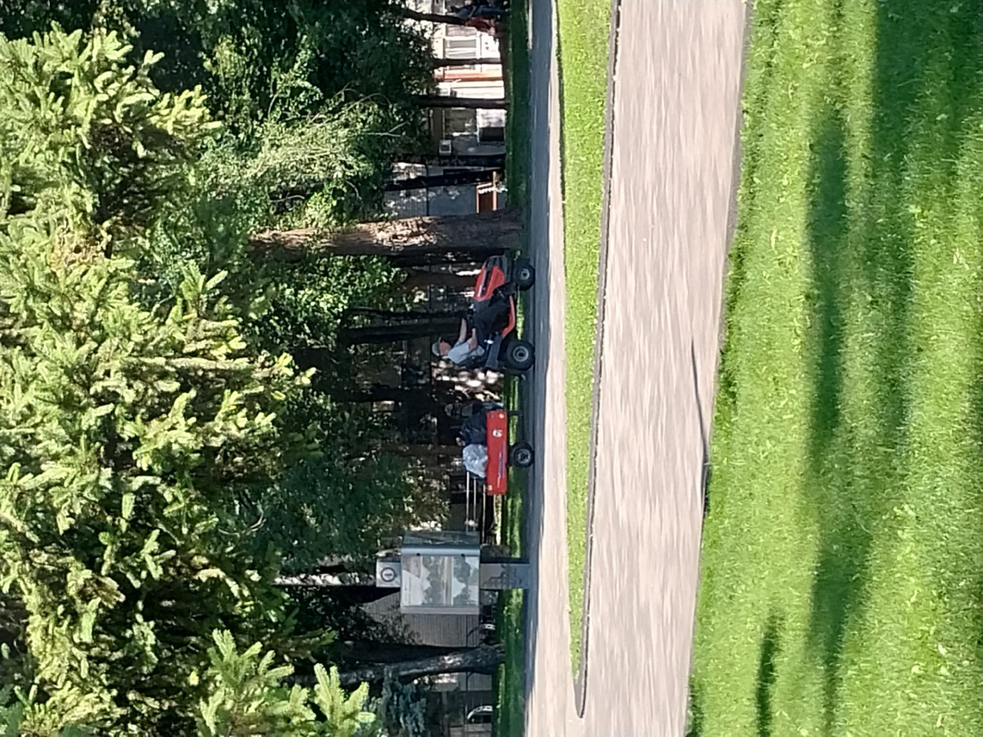 In Dnipro, Ukraine, a man is operating a lawnmower, towing a red trailer filled with bagged trash. The trailer is behind him, and he appears to be moving from right to left. In the background, there are trees, buildings, and a paved walkway. The scene suggests an outdoor maintenance activity.