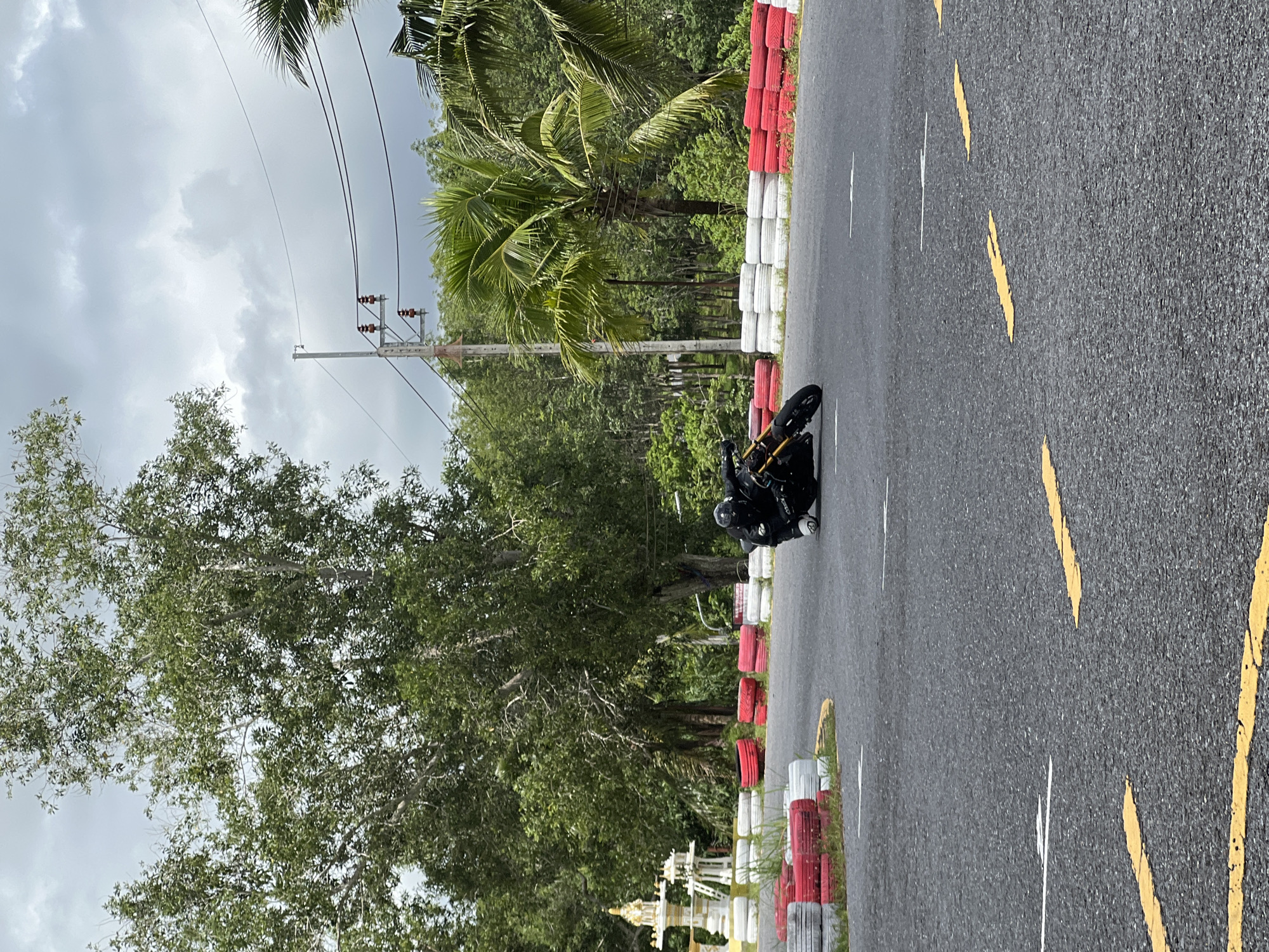 In Thep Krasatti, Thailand, a motorcycle rider wearing a black suit and helmet is leaning into a turn on an asphalt track. The track is bordered by stacks of red and white tires, with some overgrown grass along the edges. In the background, lush green trees and palm trees form a dense backdrop, with a power line pole visible in the distance under a cloudy sky. To the left, a small, ornate white structure, possibly a shrine, sits near the track. The road surface has yellow and white markings indicating lanes and boundaries.